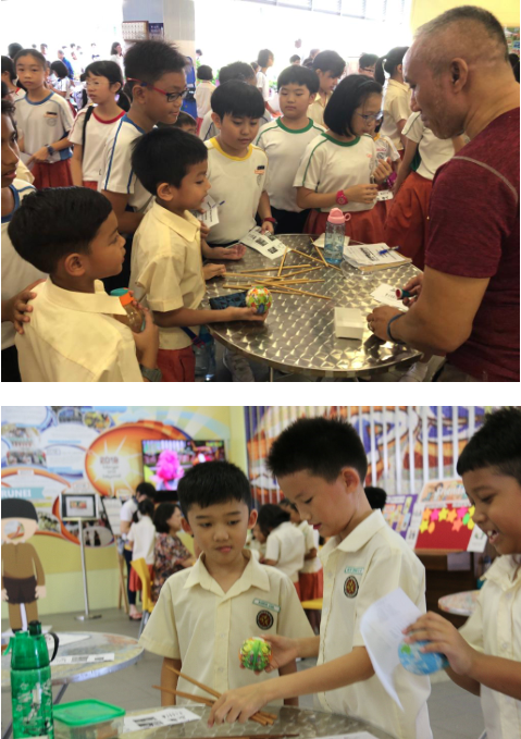 Children in school uniforms gather around tables with science fair exhibits, interacting with displays and an adult.
