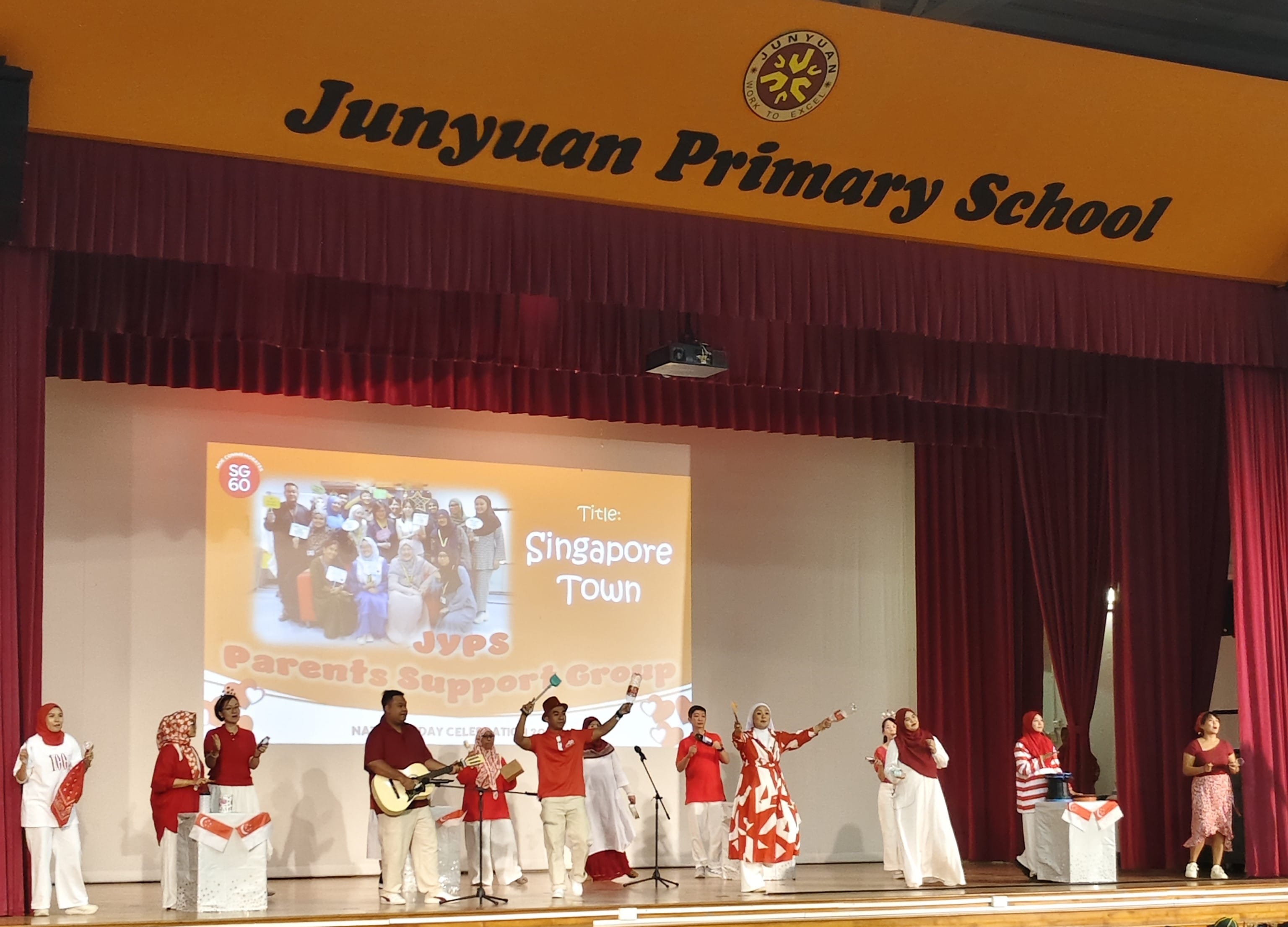 A group in red and white performs on Junyuan Primary School stage with a Singapore Town National Day slide projected behind them.