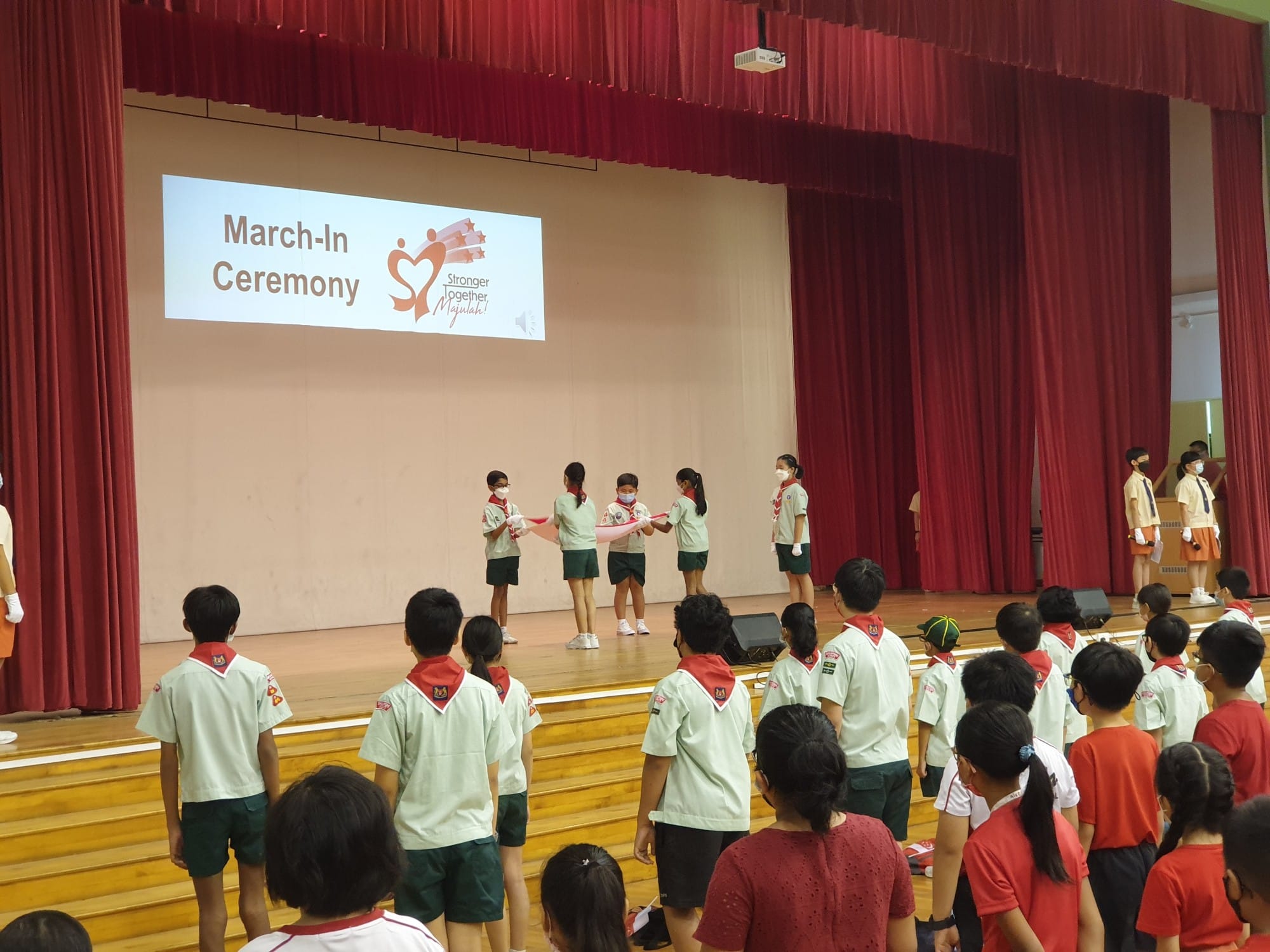 Children in scout uniforms stand in a hall during a March-In Ceremony with a projected slide on a red-curtained stage.