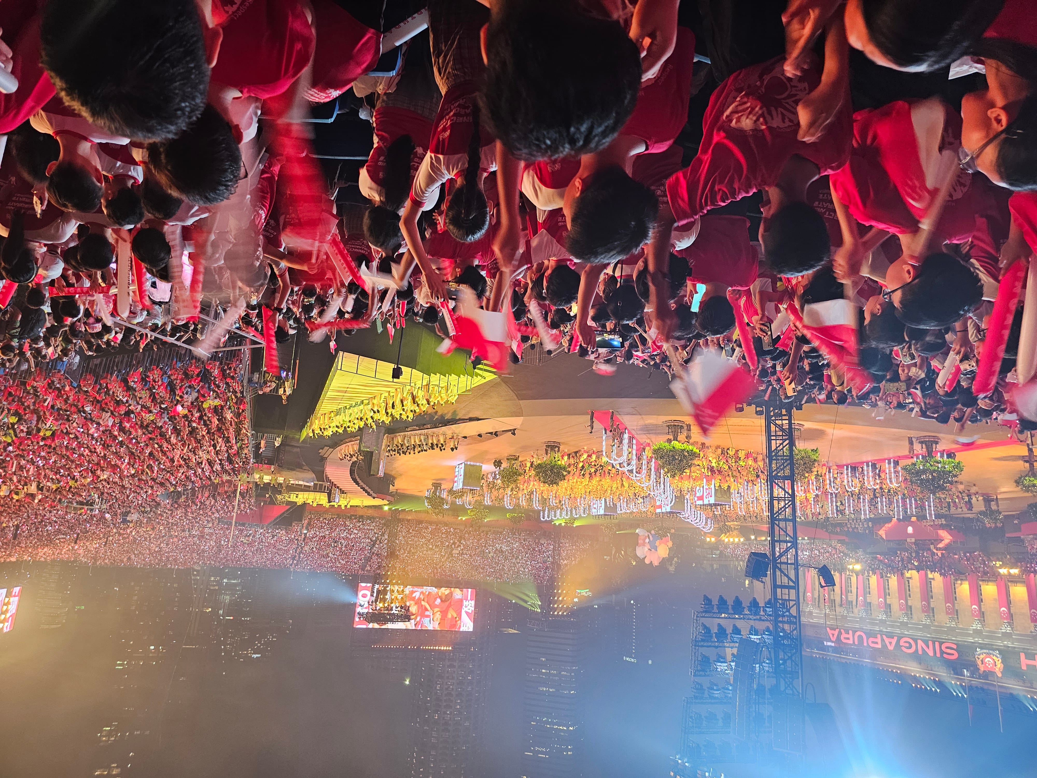 Crowd in red shirts waving small red-and-white flags at a nighttime stadium event with bright lights.