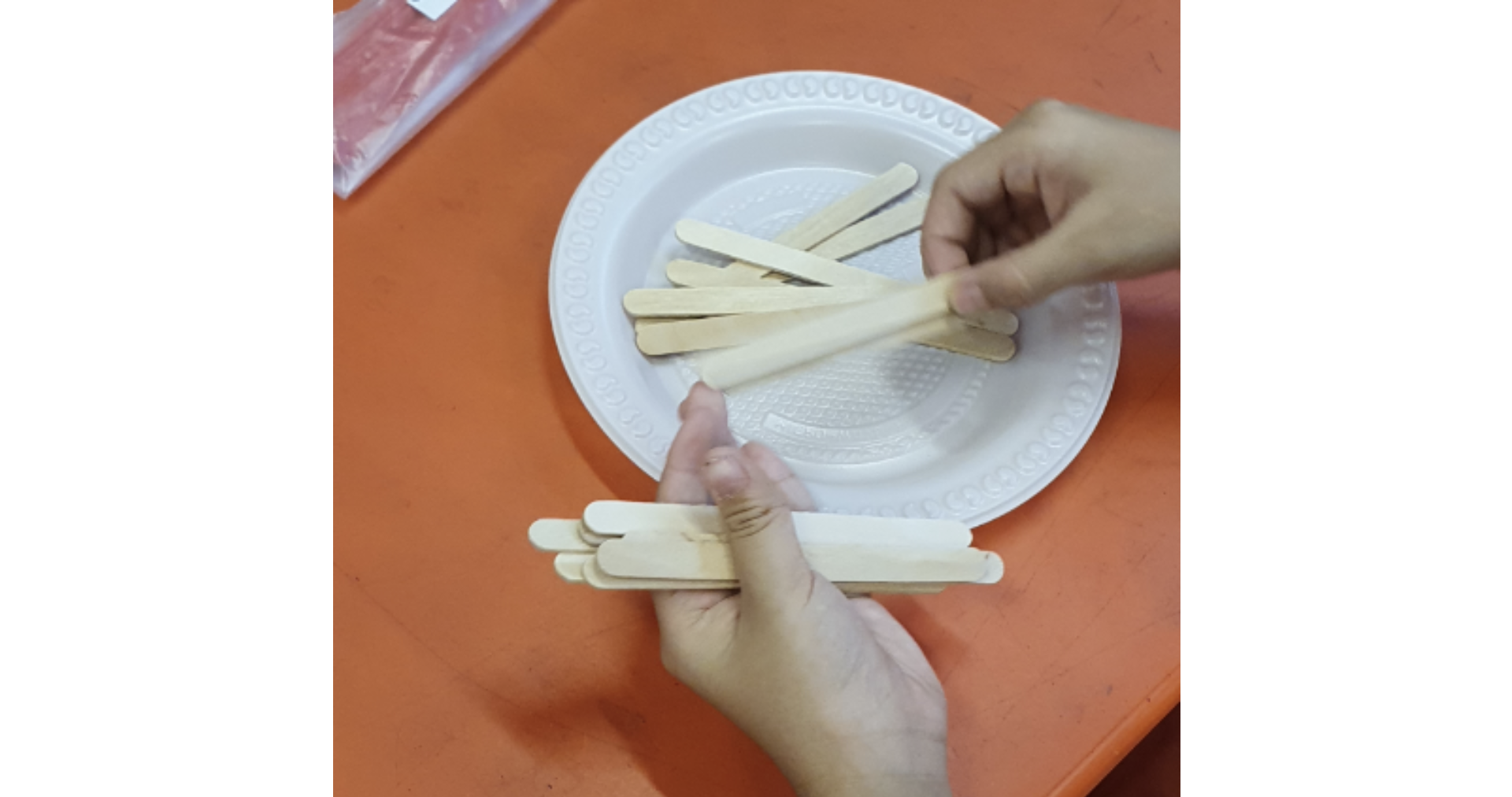 Two hands arranging wooden craft sticks on a white plate and holding a small bundle on an orange table.