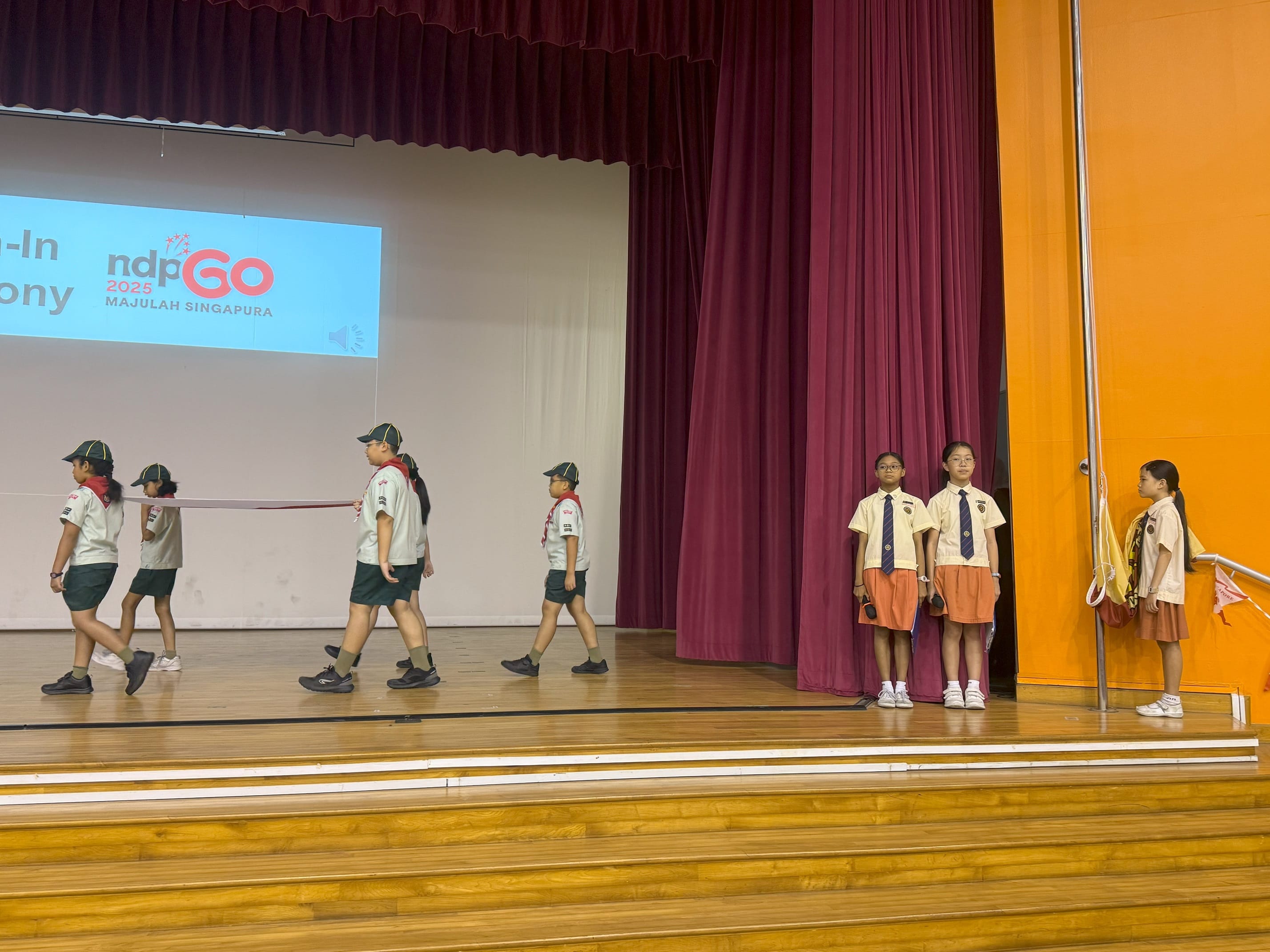 Students in uniforms march on a school stage during an NDP 2025 Majulah Singapura flag-in ceremony.