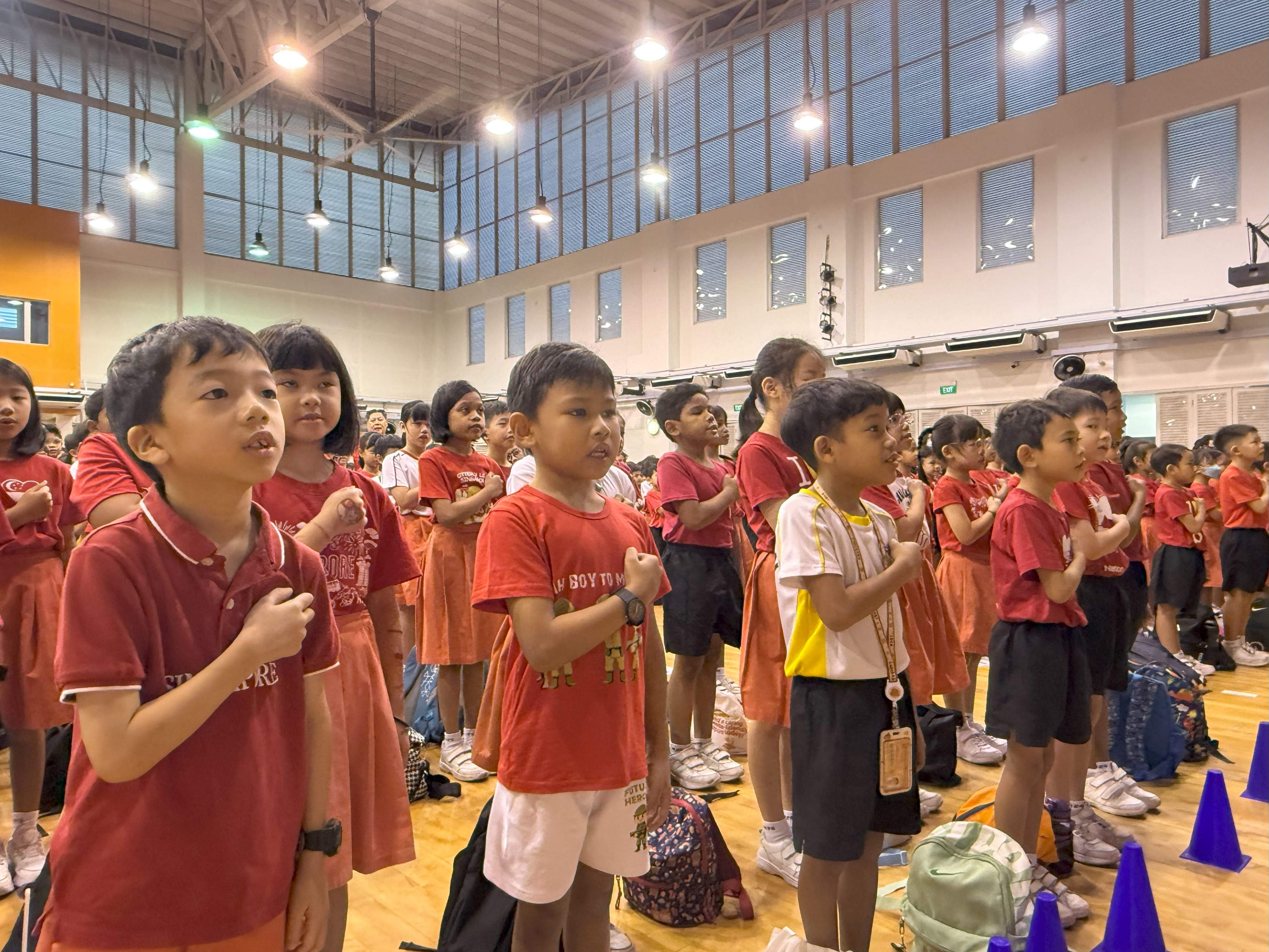 A large group of young children in red shirts stand with fists to chests in a school gymnasium.