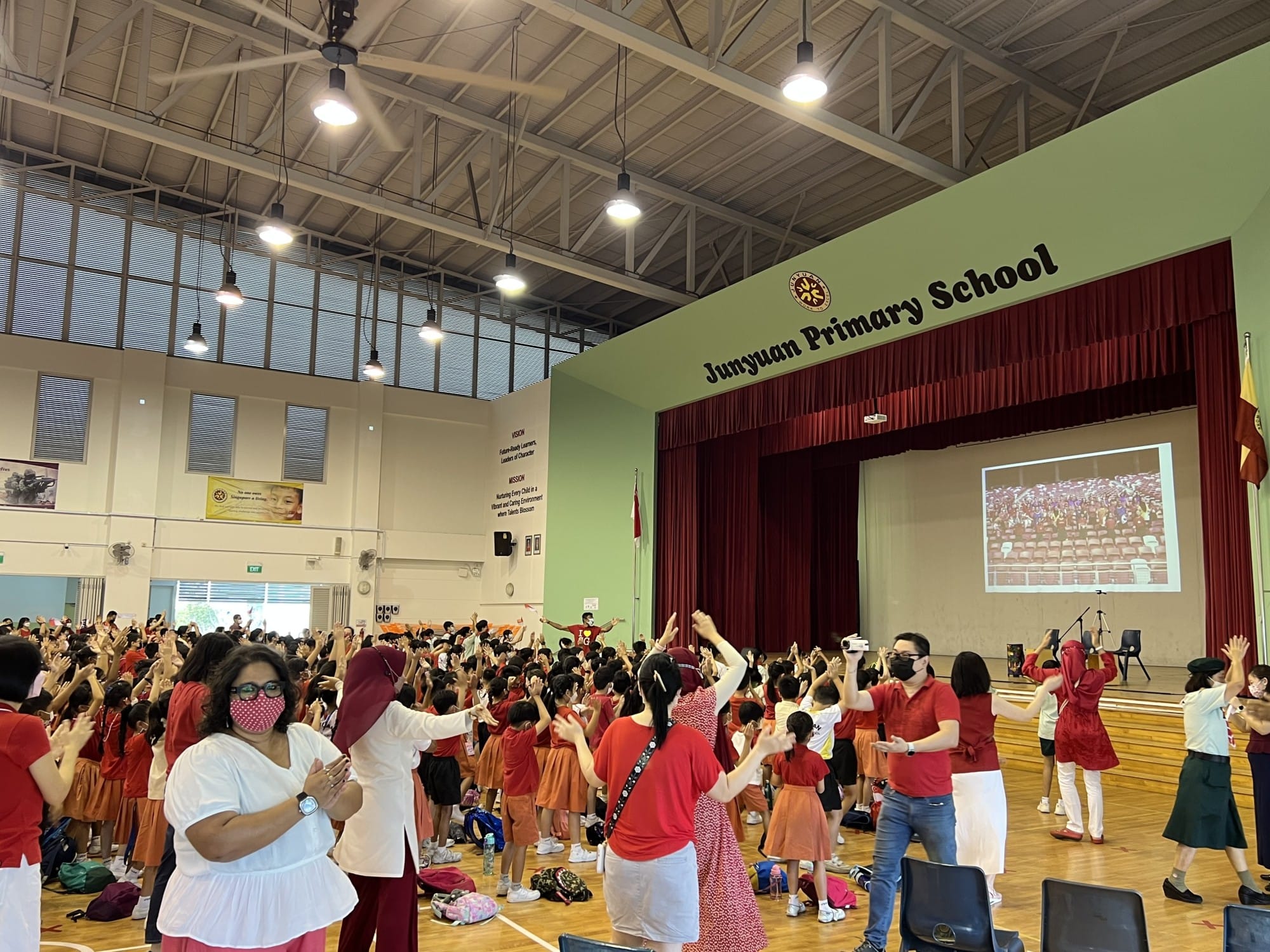 Students and teachers in red outfits dance with raised arms in Junyuan Primary School's indoor hall.