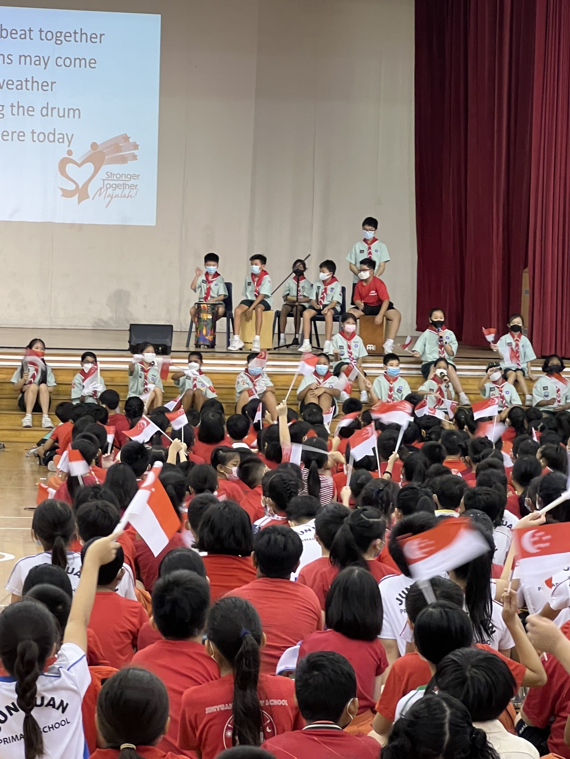 Students in red uniforms wave Singapore flags at a school assembly, with masked scouts on stage and a projected screen.