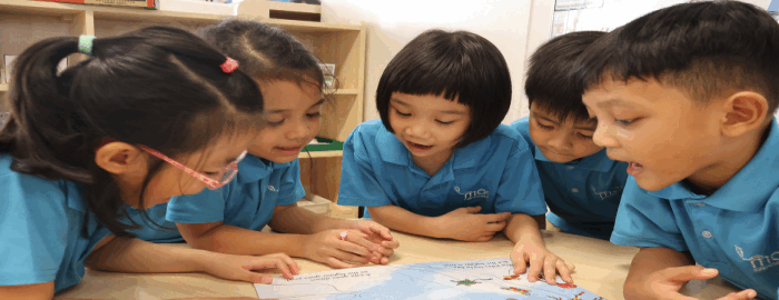Five young children in matching blue uniforms lean over a table, reading and discussing a colorful book together.