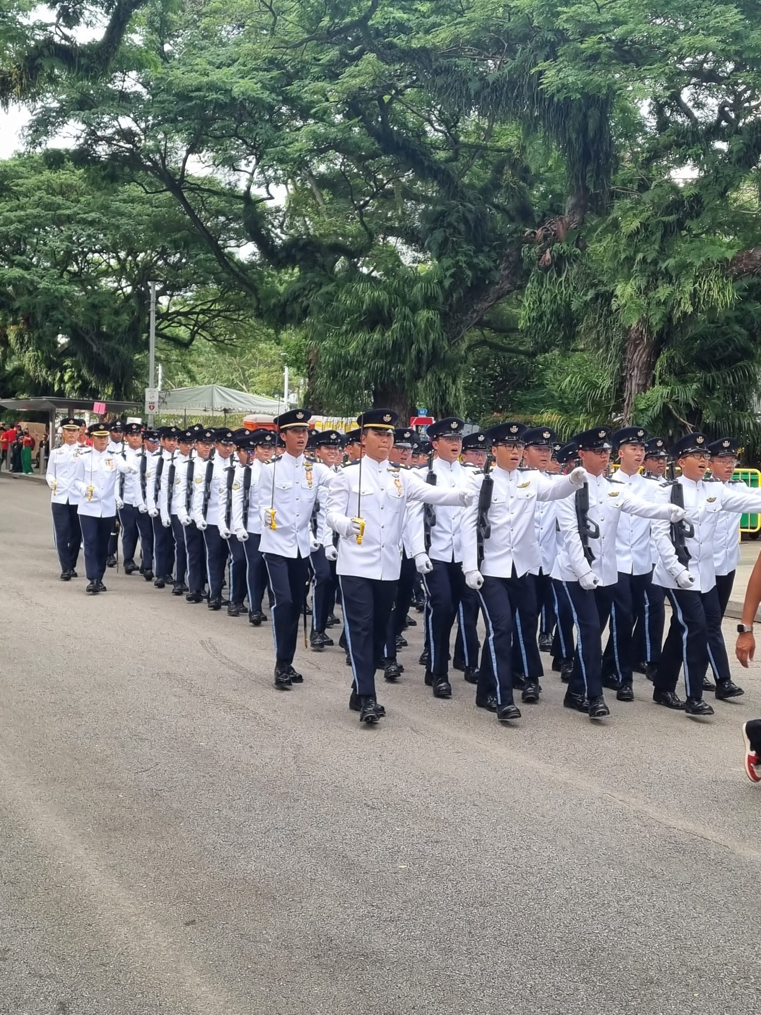 Rows of uniformed officers in white jackets, dark trousers, and black caps marching in formation on a street.