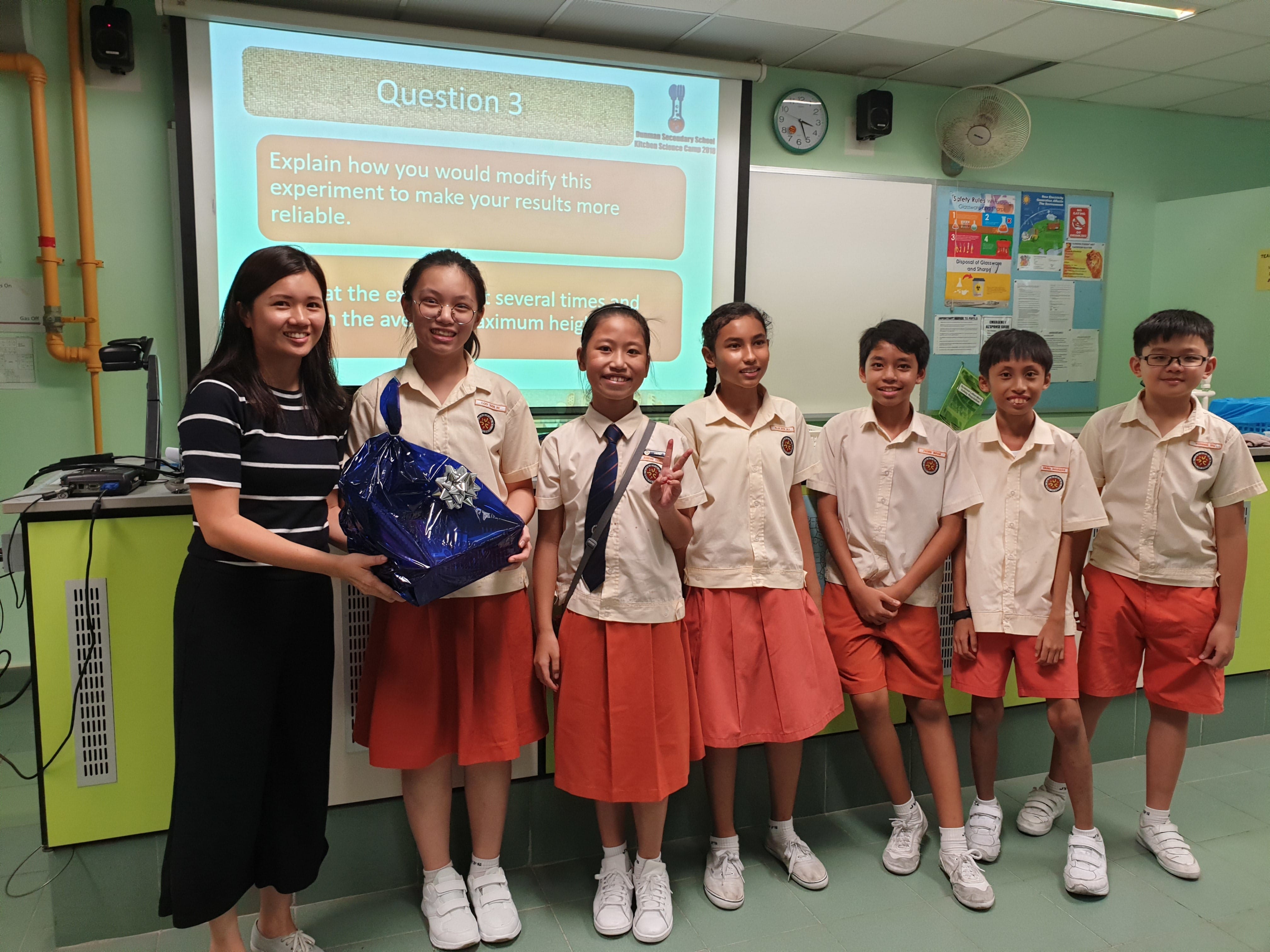 A teacher and six uniformed students pose smiling in a classroom, with a blue gift bag, before a projected quiz slide.
