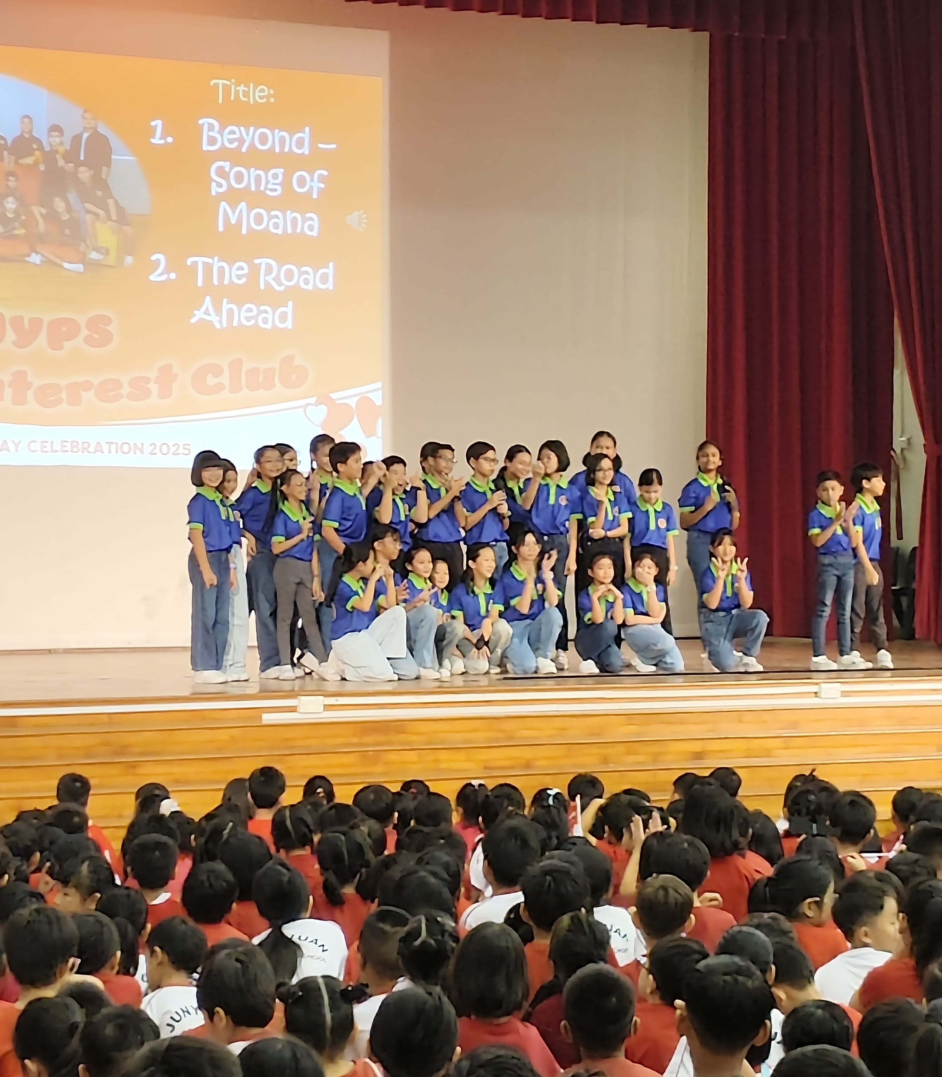 Students in blue uniforms perform on stage before a large seated audience at a 2025 school celebration event.