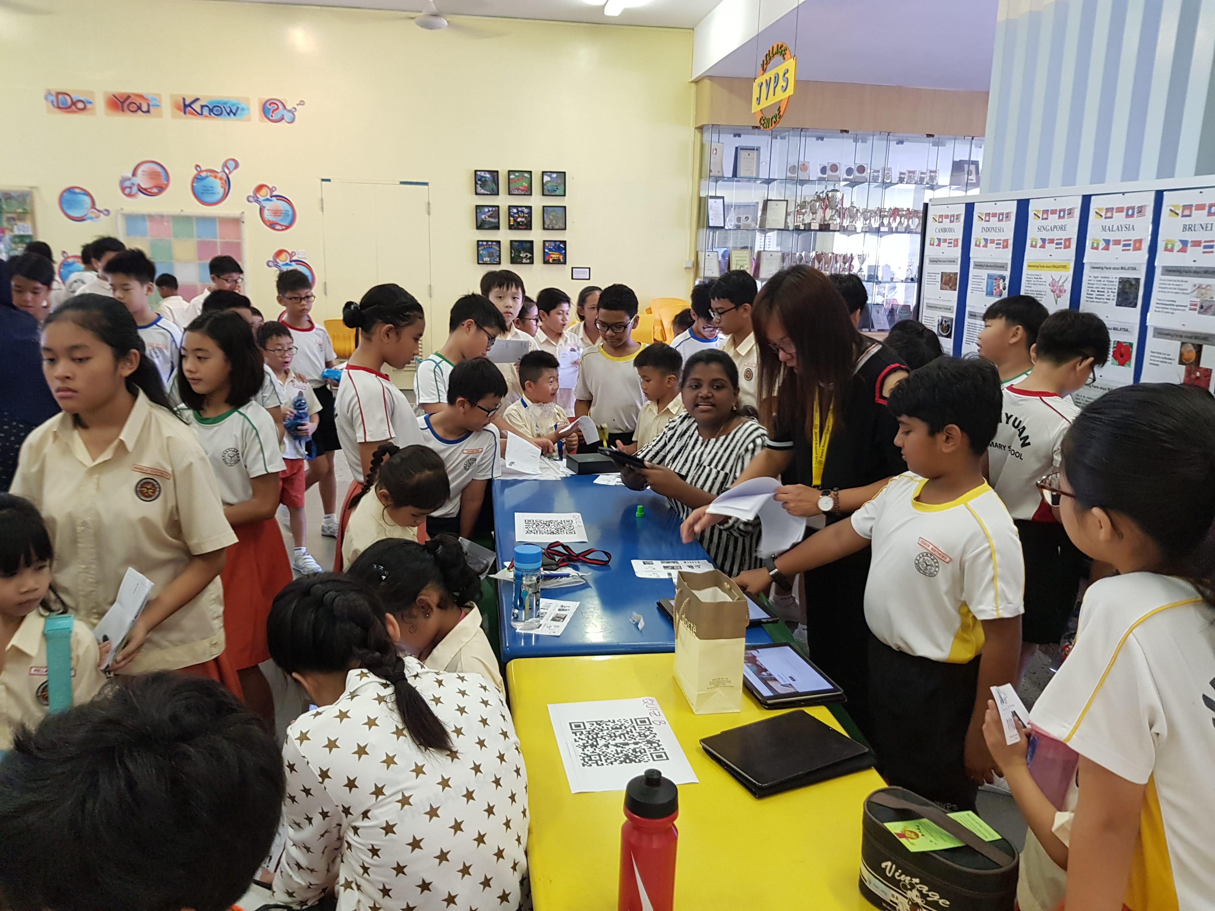 Crowd of schoolchildren around tables with QR codes, as a teacher hands out papers in a classroom.