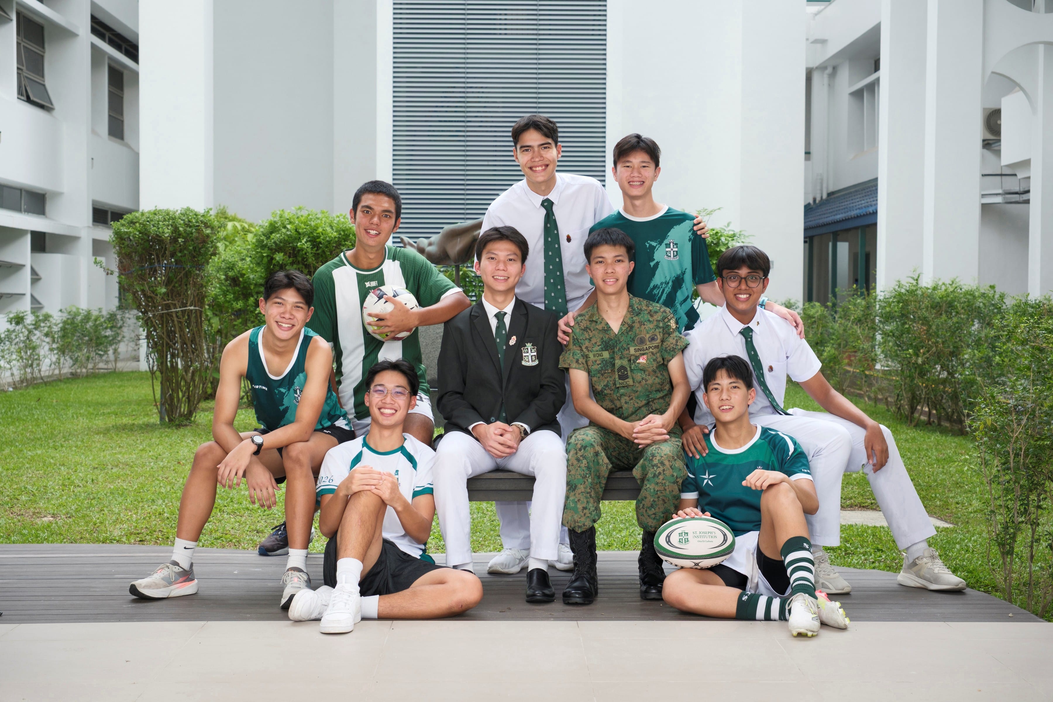 Nine young men in various uniforms and sports attire pose together outdoors, some holding a football and rugby ball.