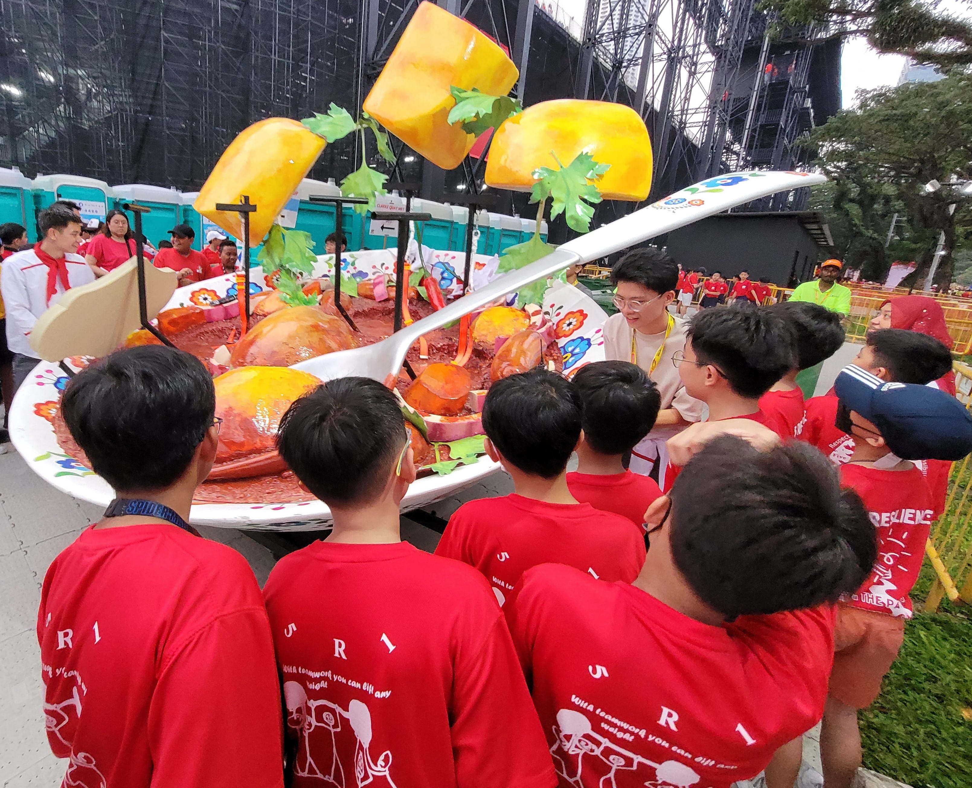 Children in red shirts gather around a large colorful parade float shaped like a giant bowl of fruit with oversized spoons.