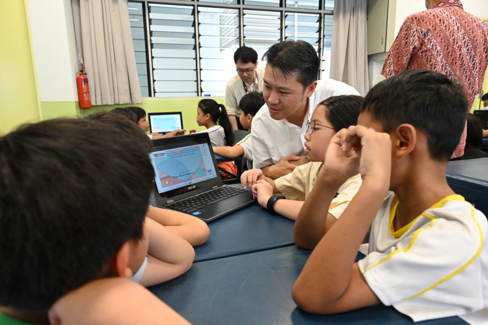 An adult leans over to guide young students looking at a laptop screen in a classroom setting.
