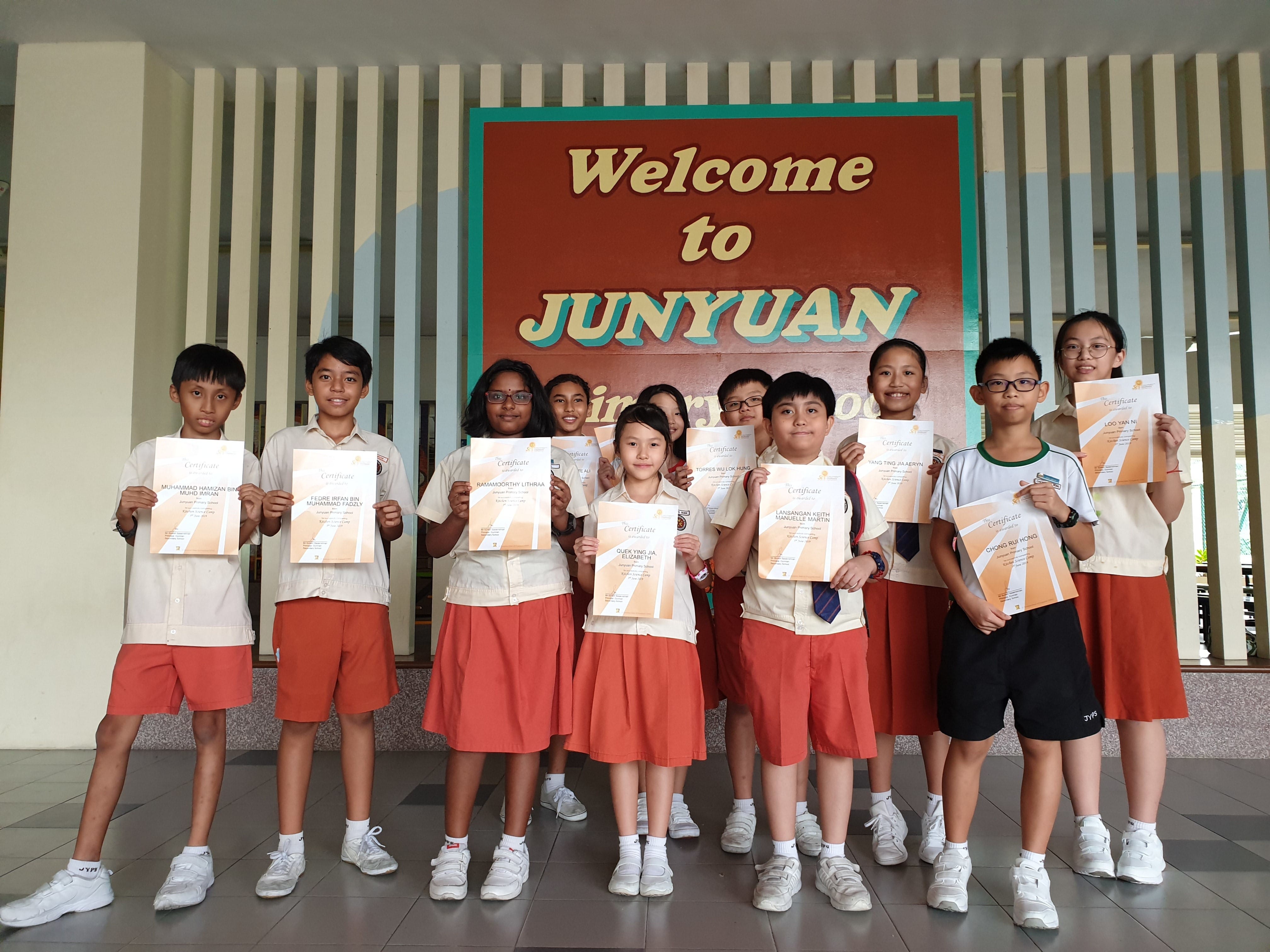 Students in uniform hold certificates in front of a Welcome to Junyuan Primary School sign.