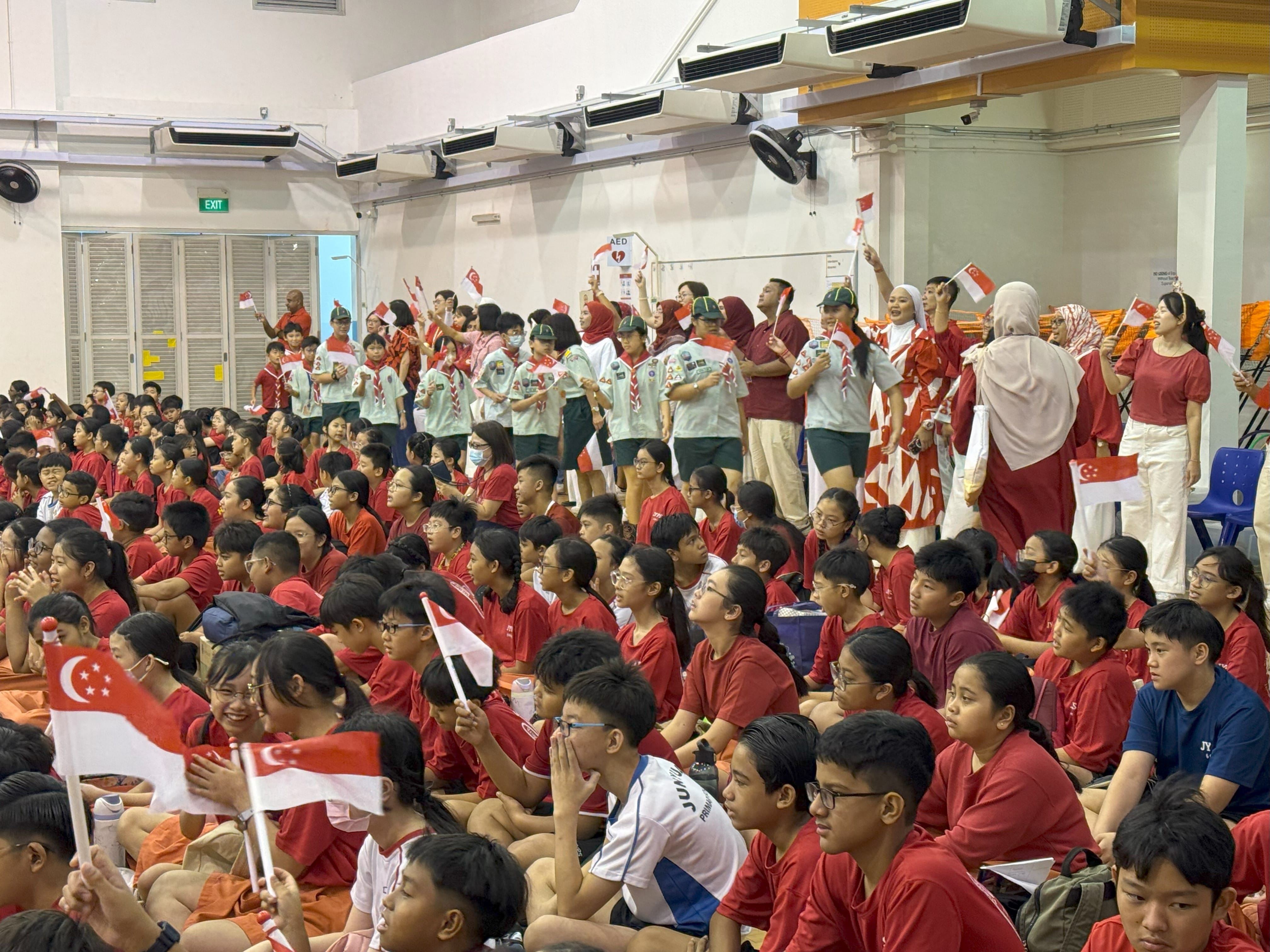 Students in red shirts sit and stand indoors waving small Singapore flags during a school celebration event.