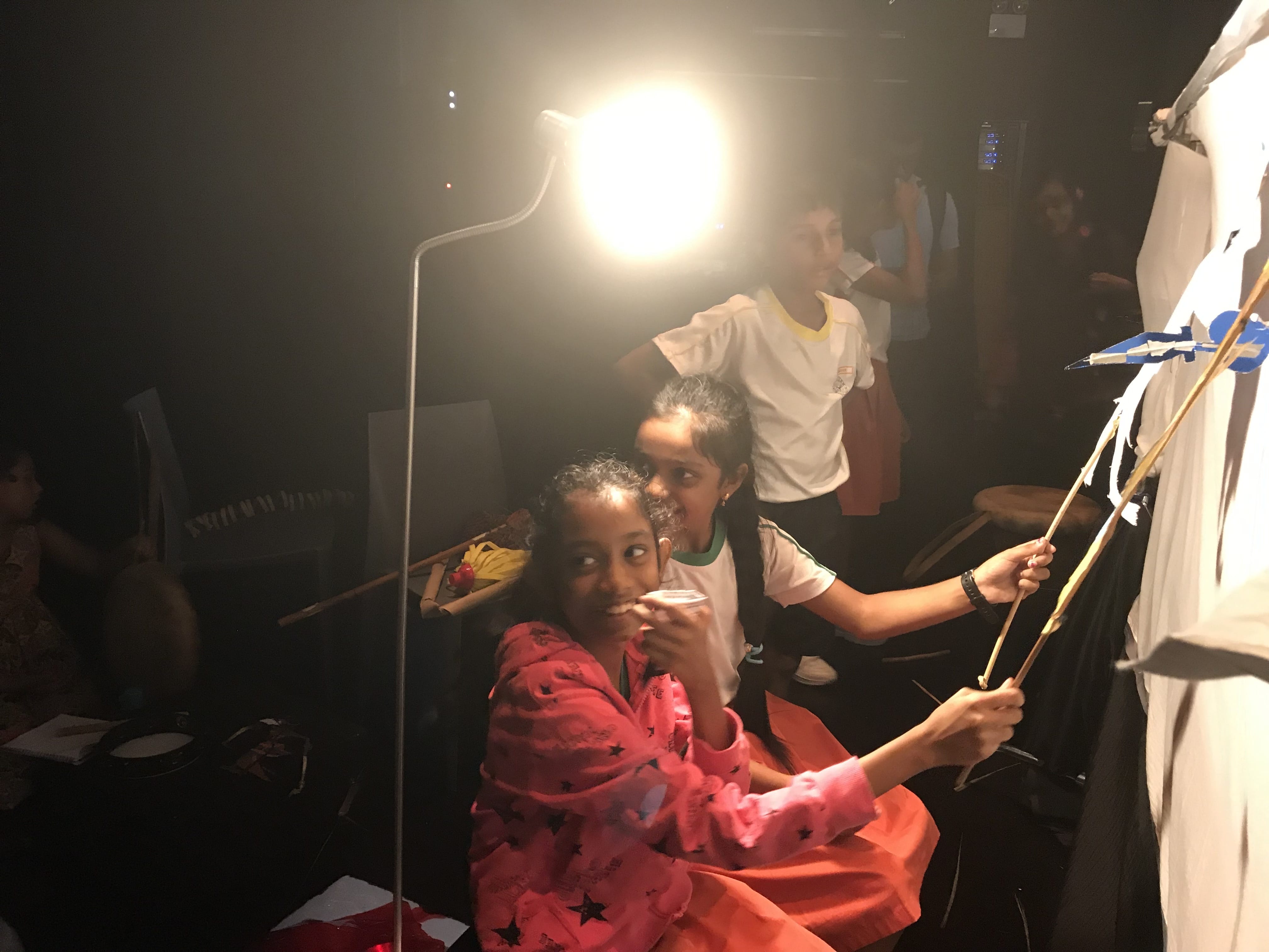 Three children in a dark room with a bright lamp, holding sticks with paper puppets near a white screen.