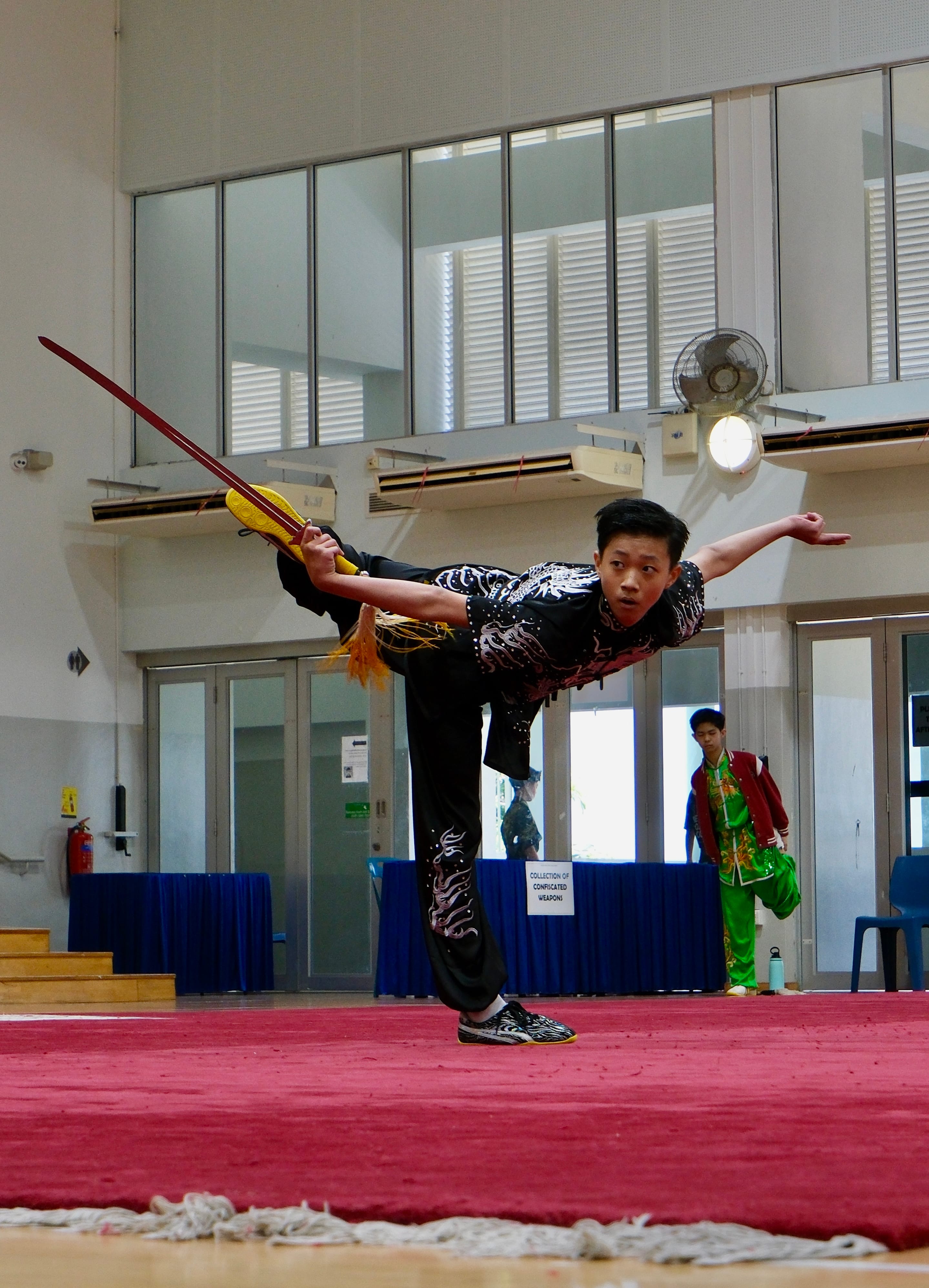 Four young practitioners in colorful wushu uniforms pose with weapons outdoors near a stone with Chinese characters.