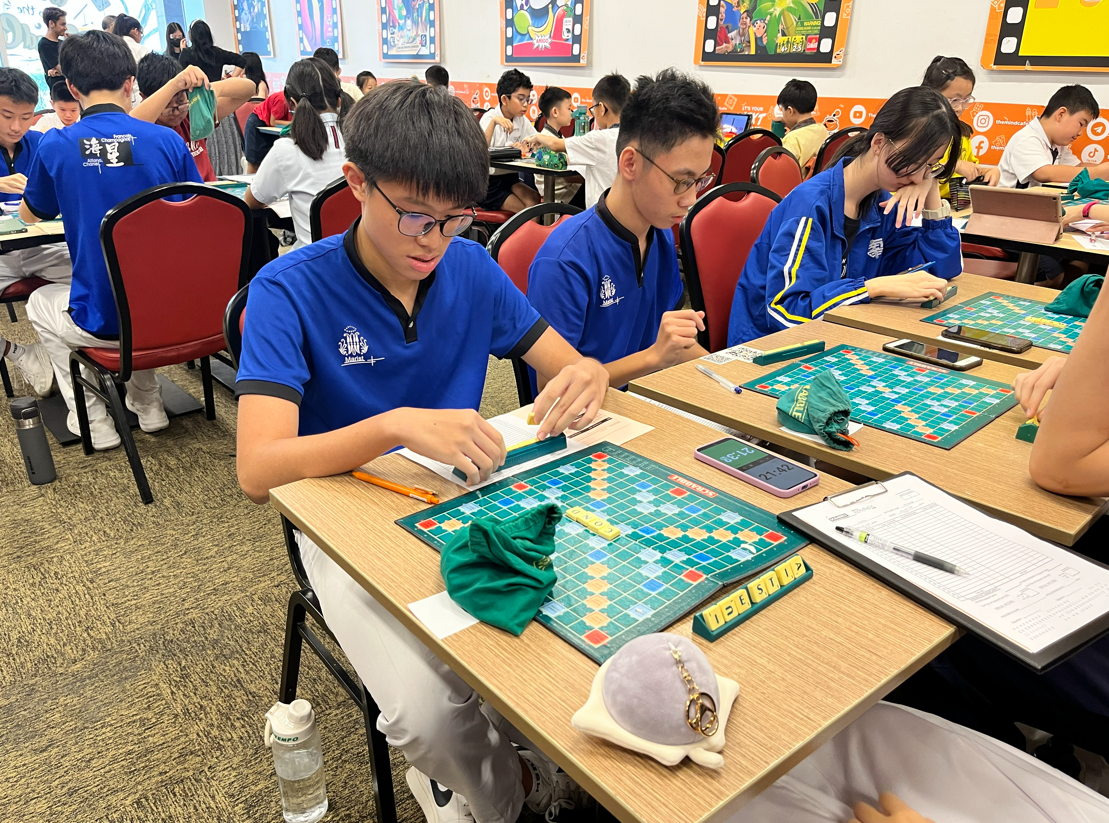 Two masked students sit across from each other at an outdoor table, each with a laptop showing an online Scrabble game.