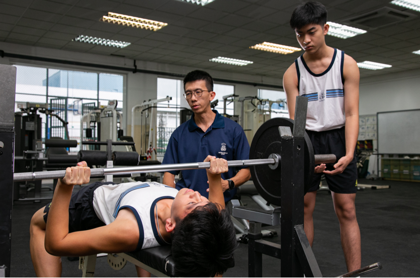 A trainer crouches beside a person lying on a bench pressing a barbell in a gym with others in the background.
