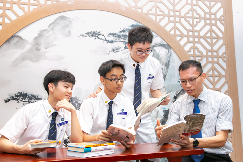 Three young men in white shirts review a booklet together at a table near a window with plants nearby.