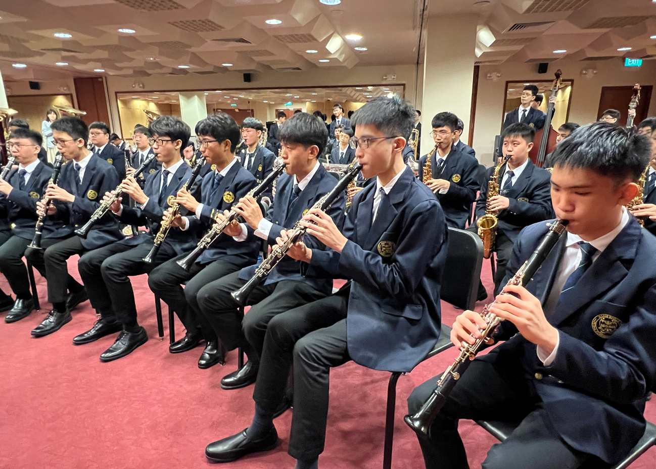 Five students in navy blazers hold brass instruments and a snare drum outdoors near palm trees and a school building.