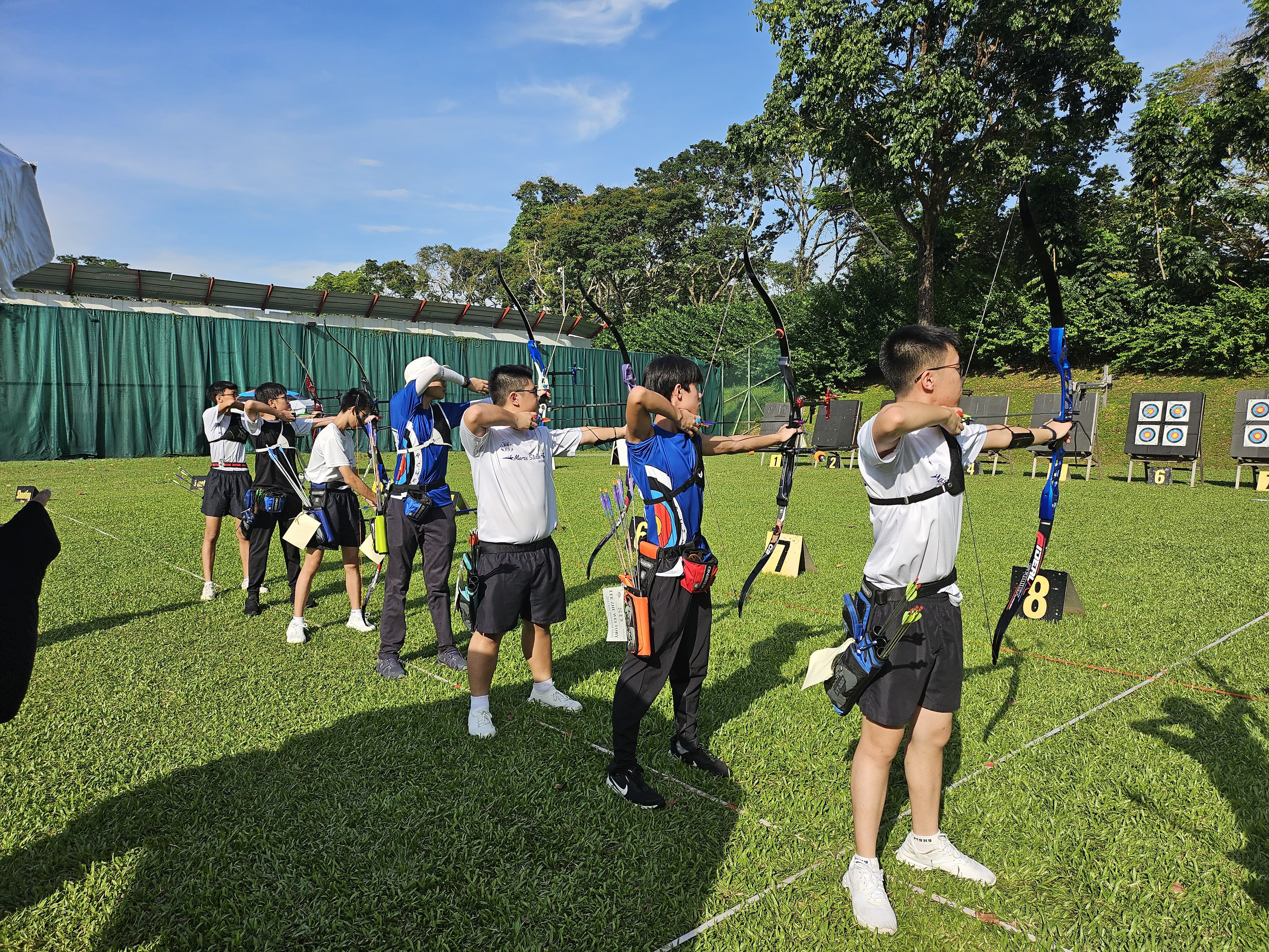 Person in a black shirt aiming a compound bow upward against a bright blue sky with sun flare.