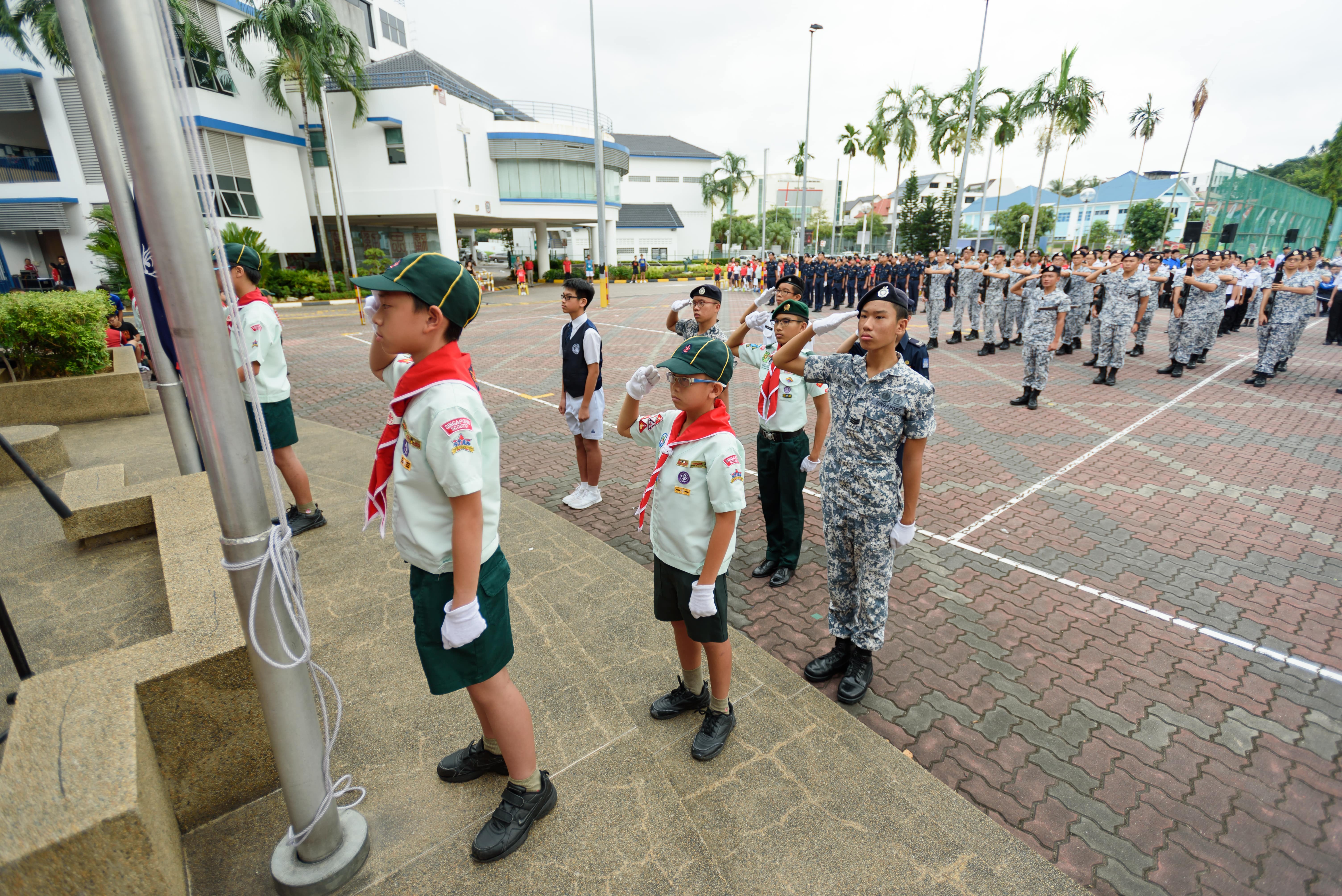 Youth scouts and uniformed cadets saluting during a flag-raising ceremony on a school parade square.