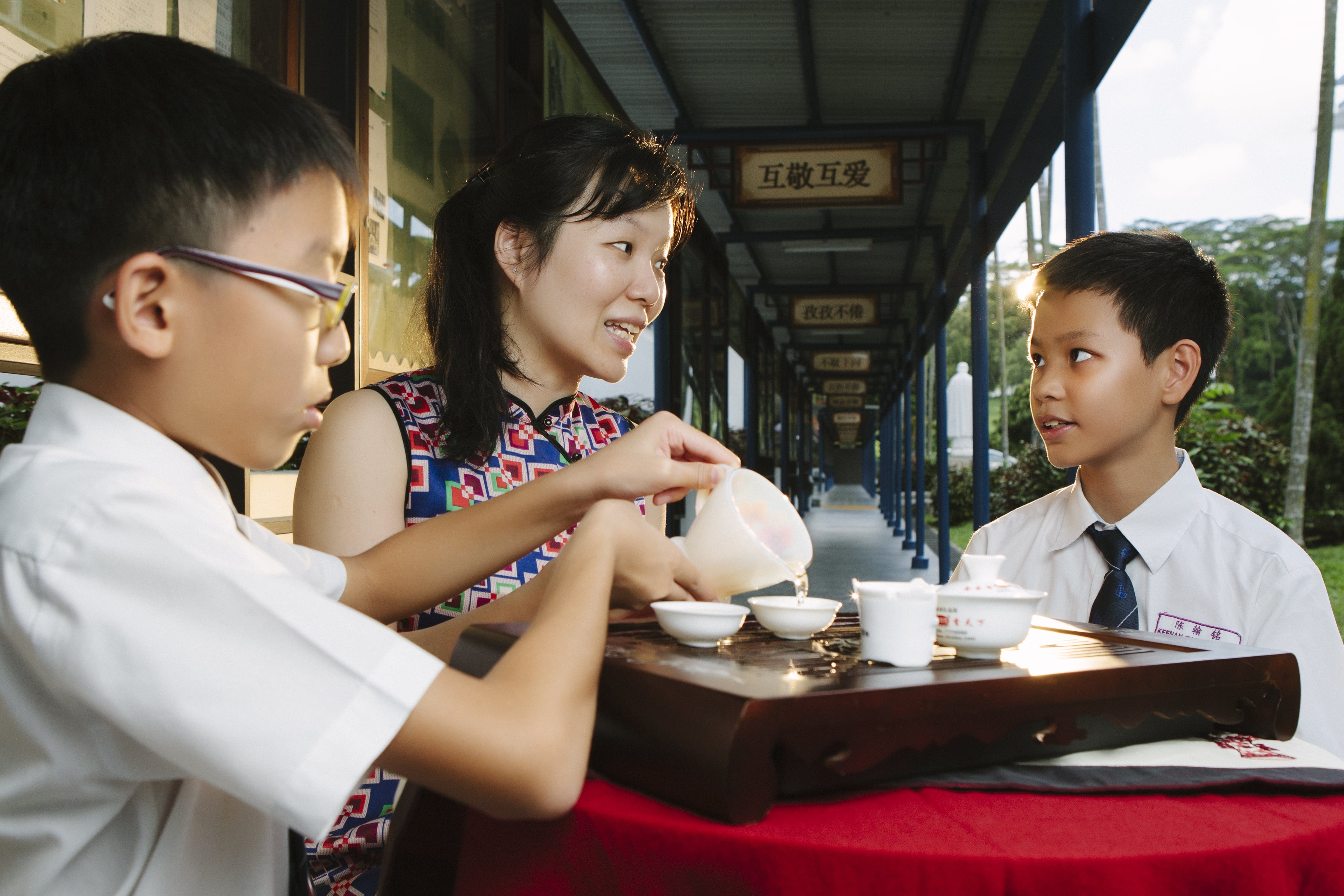 A woman in a patterned dress pours tea for two boys in school uniforms at a table in a covered walkway.