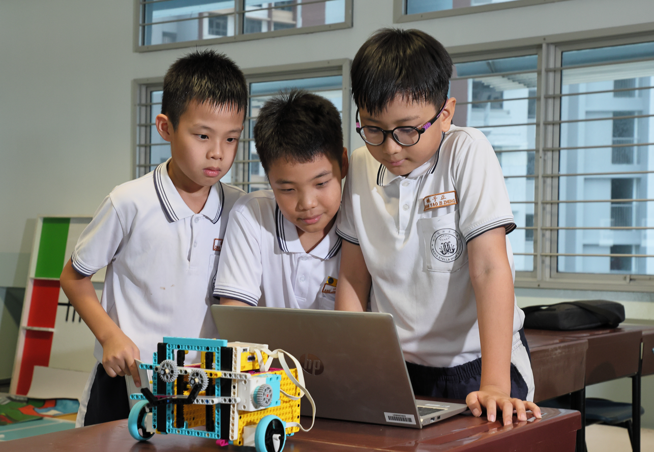 Three boys in white uniforms lean over a table, focused on building a small LEGO robot.
