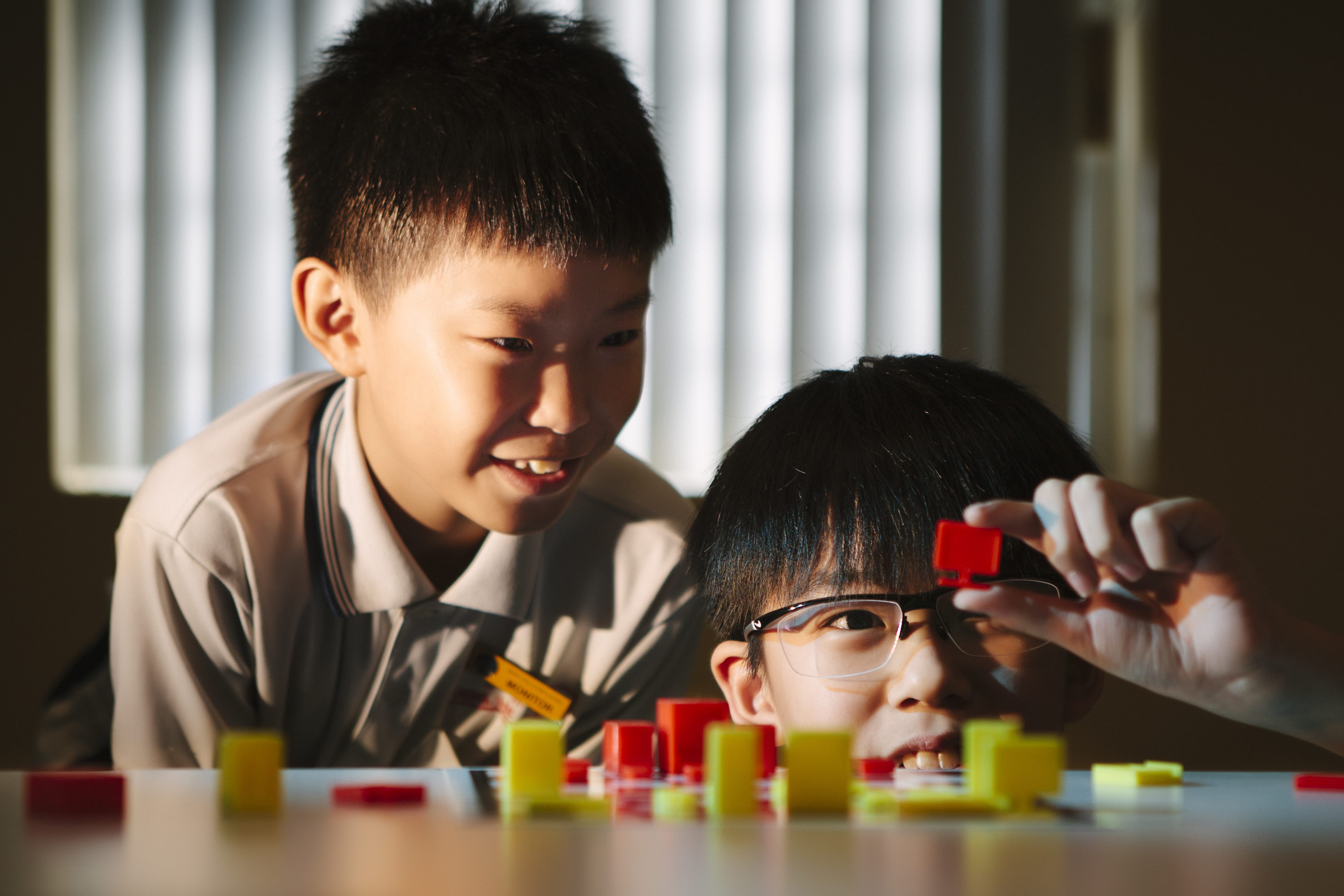 Two young boys smile while playing with red and yellow plastic cubes on a table near a window.