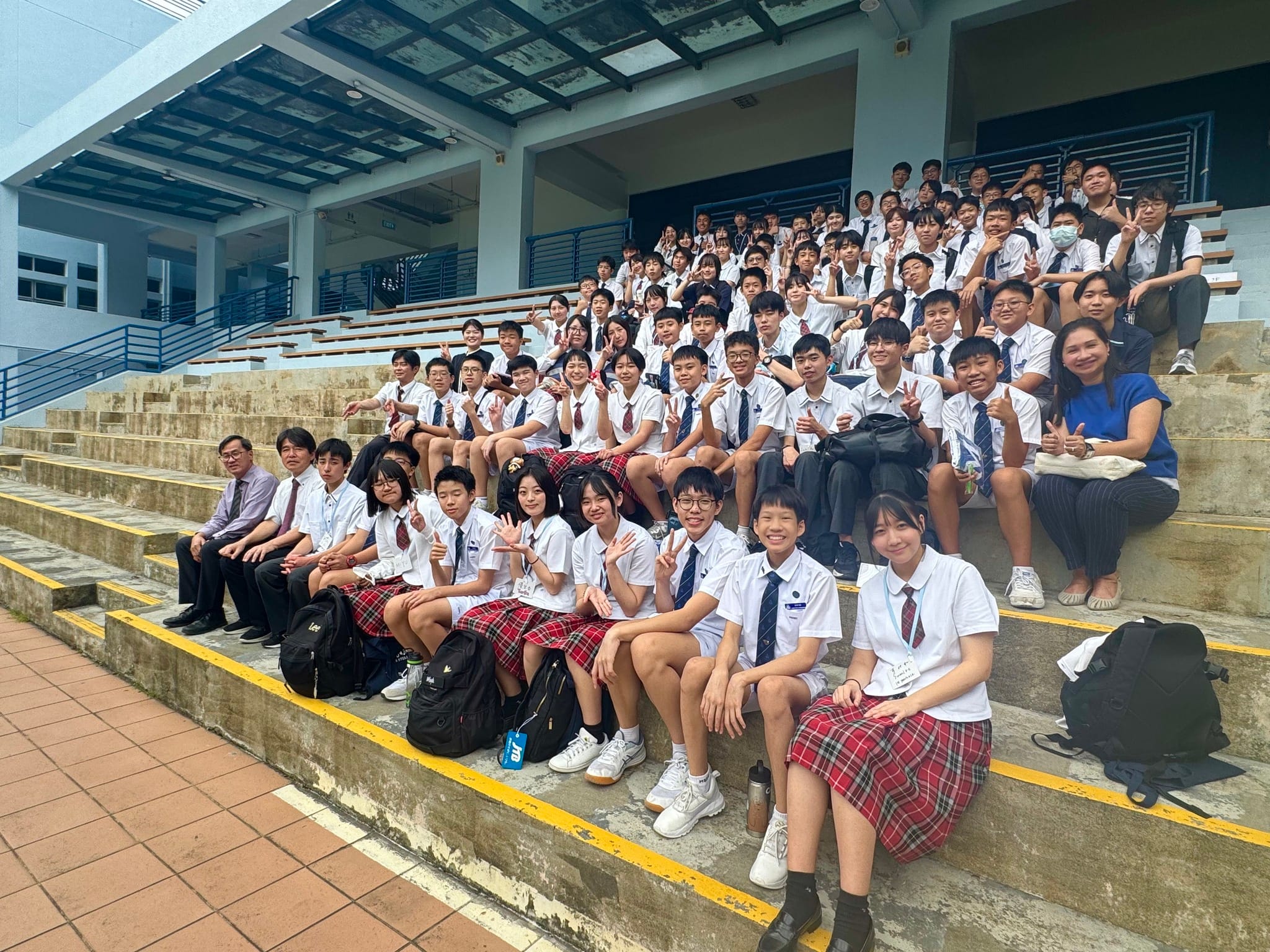 A large group of uniformed school students and teachers posing on outdoor bleacher steps, many waving and smiling.