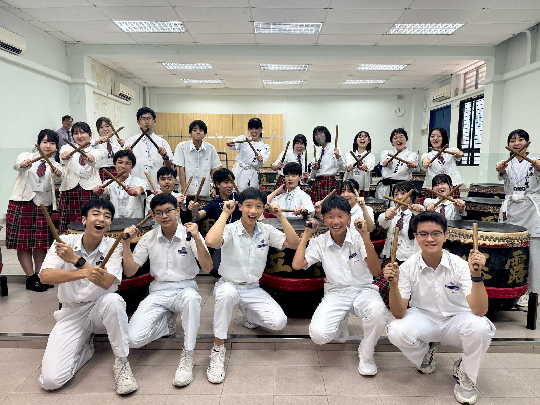 A group of students in white uniforms hold drumsticks and pose smiling around large taiko drums in a classroom.