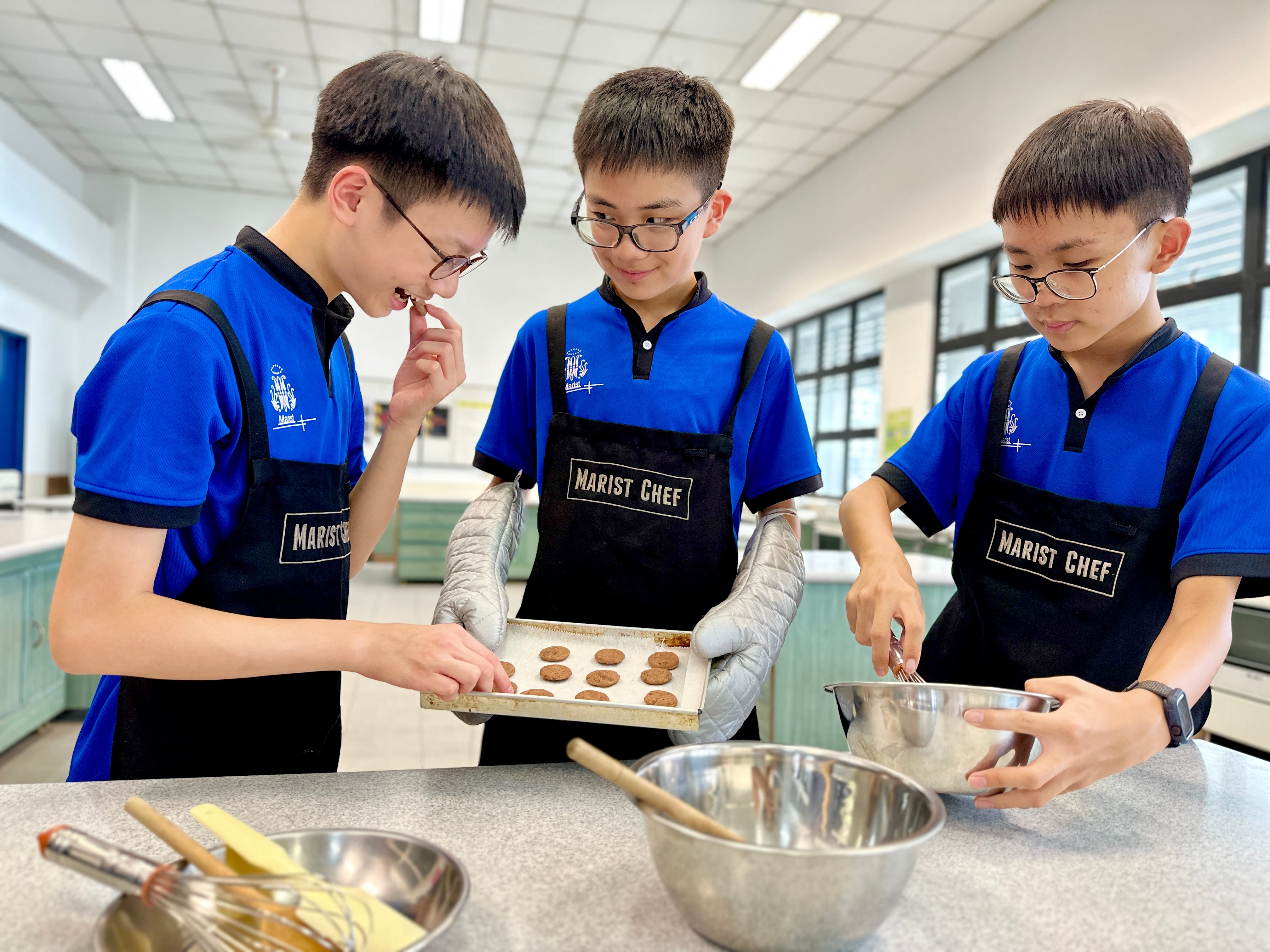 Three students in black aprons labeled Junior Chef gather around a cooking station, one reading a recipe book.