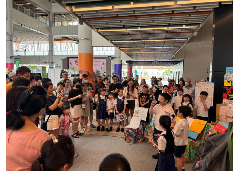 Children in school uniforms present projects to a crowd of adults and kids in a modern indoor hall.