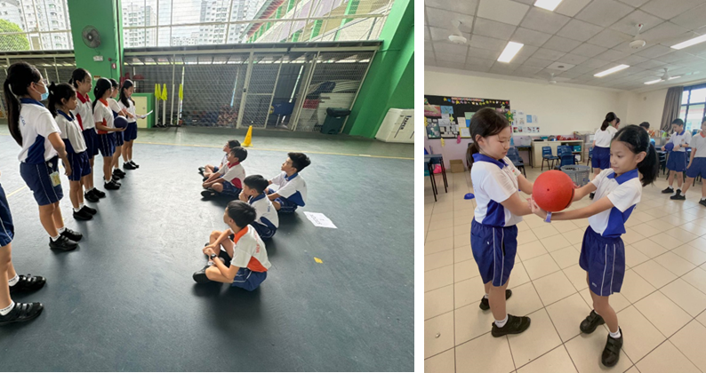Two school children in uniform pass an orange ball indoors, while other students line up or sit on a gym floor nearby.