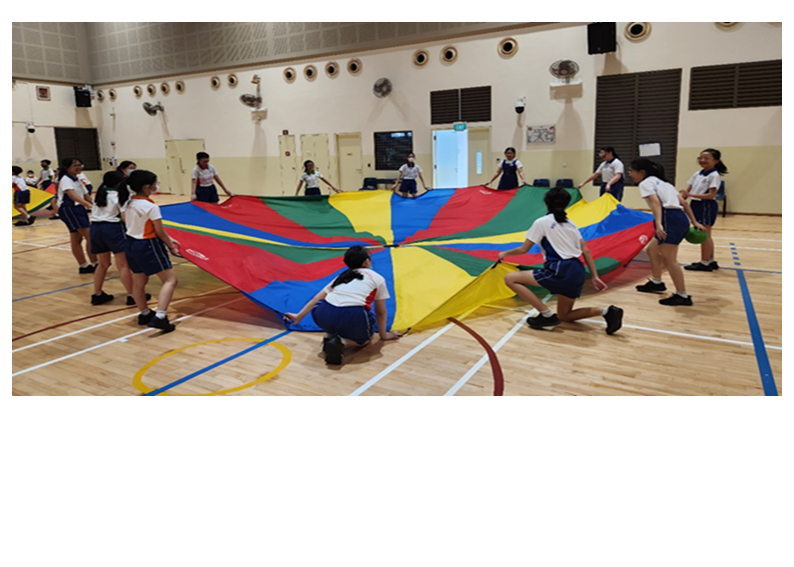 Students in school uniforms play with a large colorful parachute on an indoor gymnasium floor.