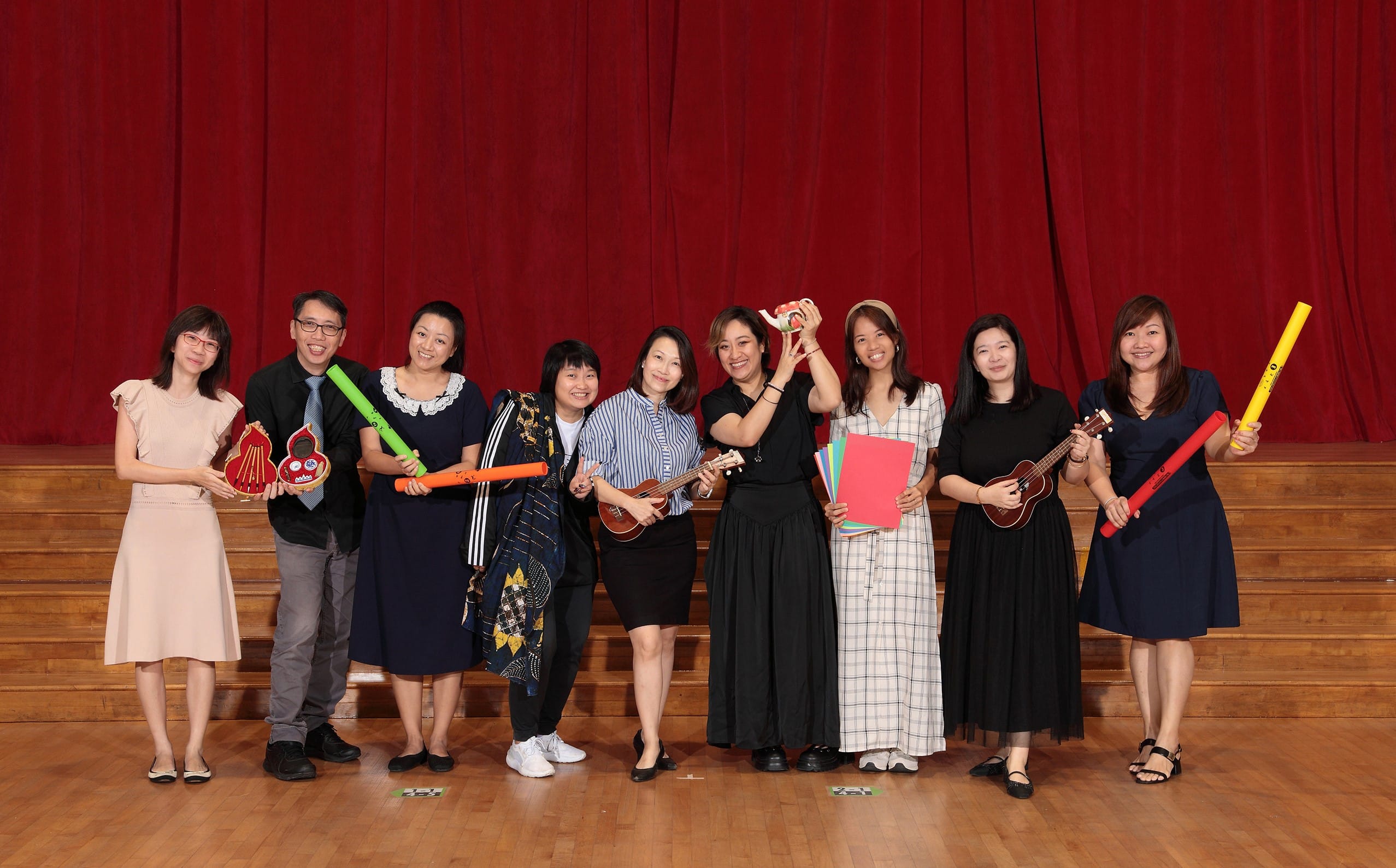 Nine teachers stand smiling in front of the wooden stage holding musical instruments and colorful items.