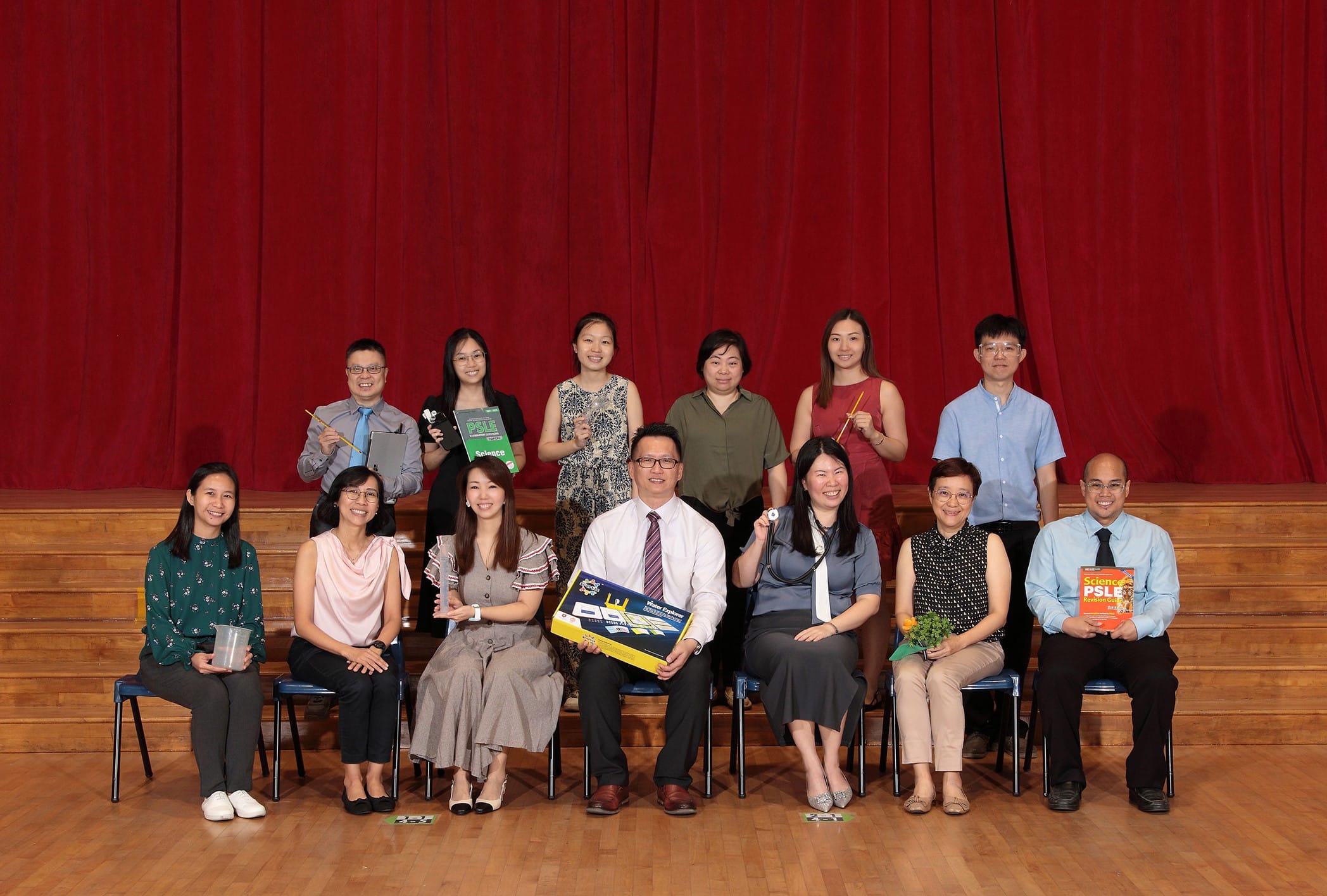 Group of thirteen adults posing on a wooden stage in front of a red curtain, some holding books and items.