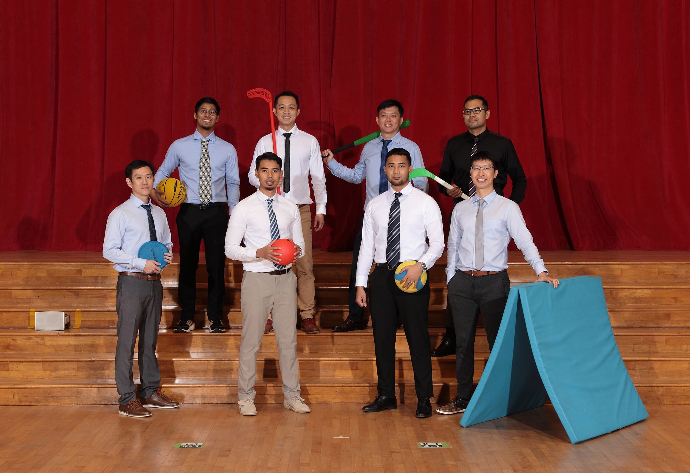 Eight men in dress shirts and ties pose on gym bleachers holding sports equipment including balls and hockey sticks.