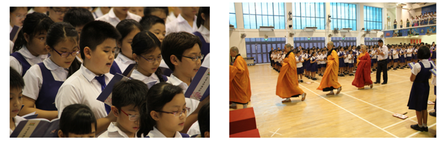 Students in white and blue uniforms sing from books while monks in orange and red robes walk through a school hall.