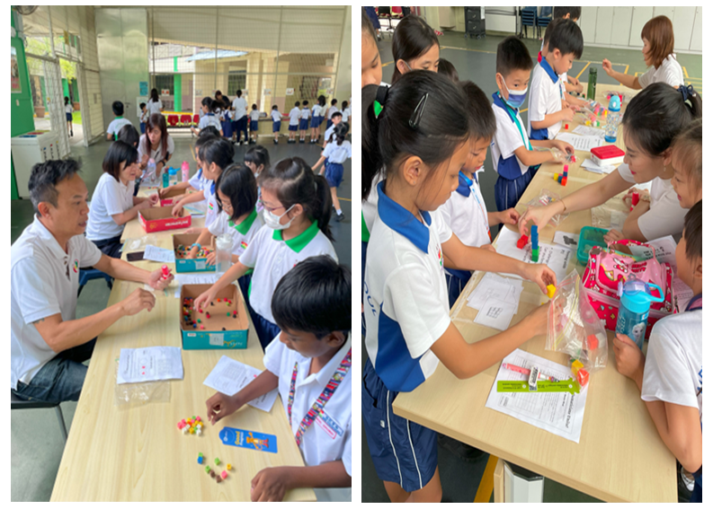 Young students in white uniforms gather around tables with colorful building pieces, guided by adults in a school setting.