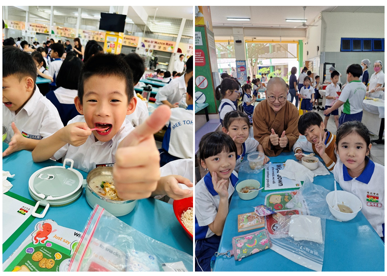 Young students eating lunch at school cafeteria tables, one boy gives a thumbs up, others pose smiling with a monk.