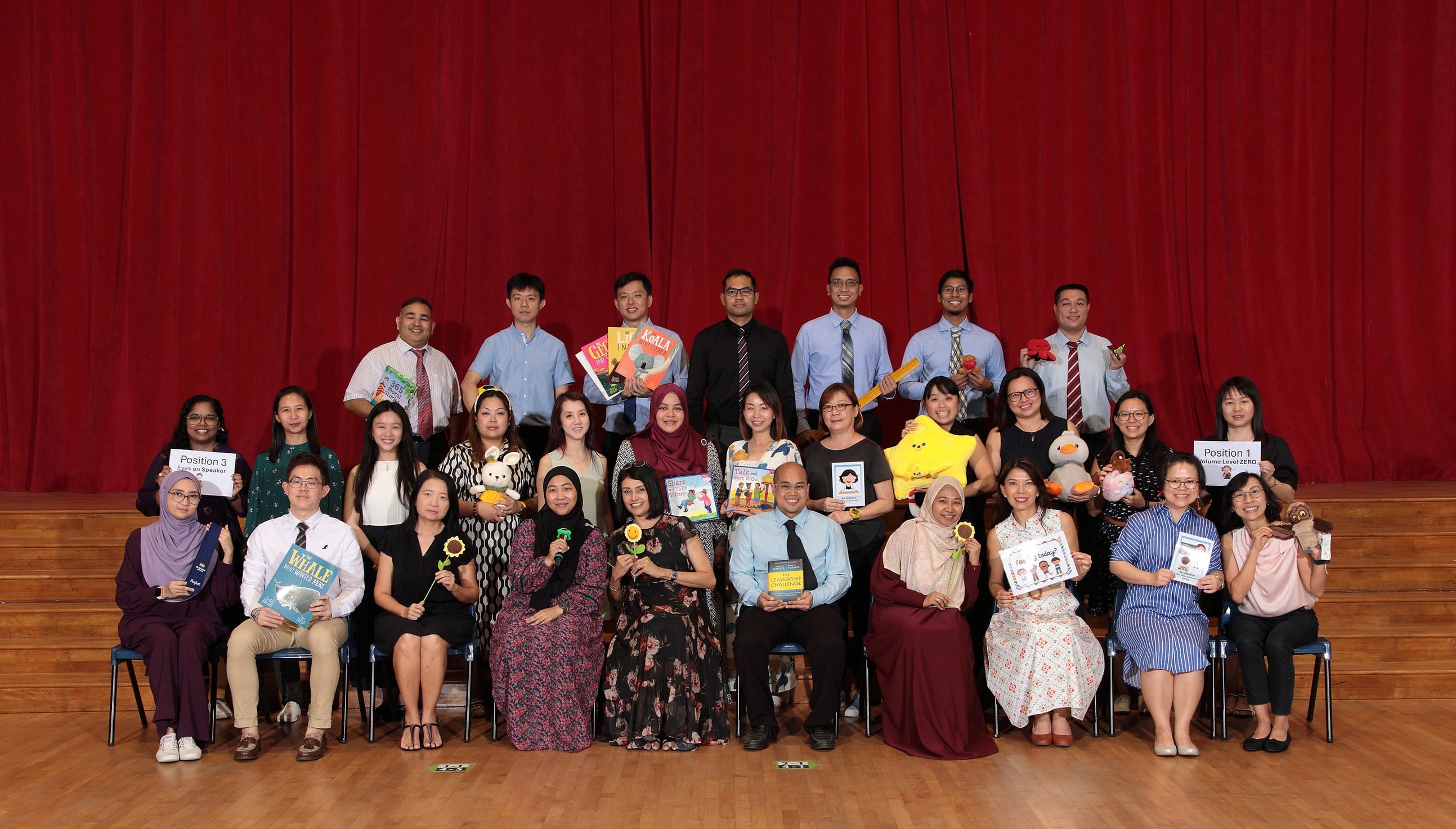 A group of teachers posing on a wooden stage before a red curtain, holding books, toys, and signs.