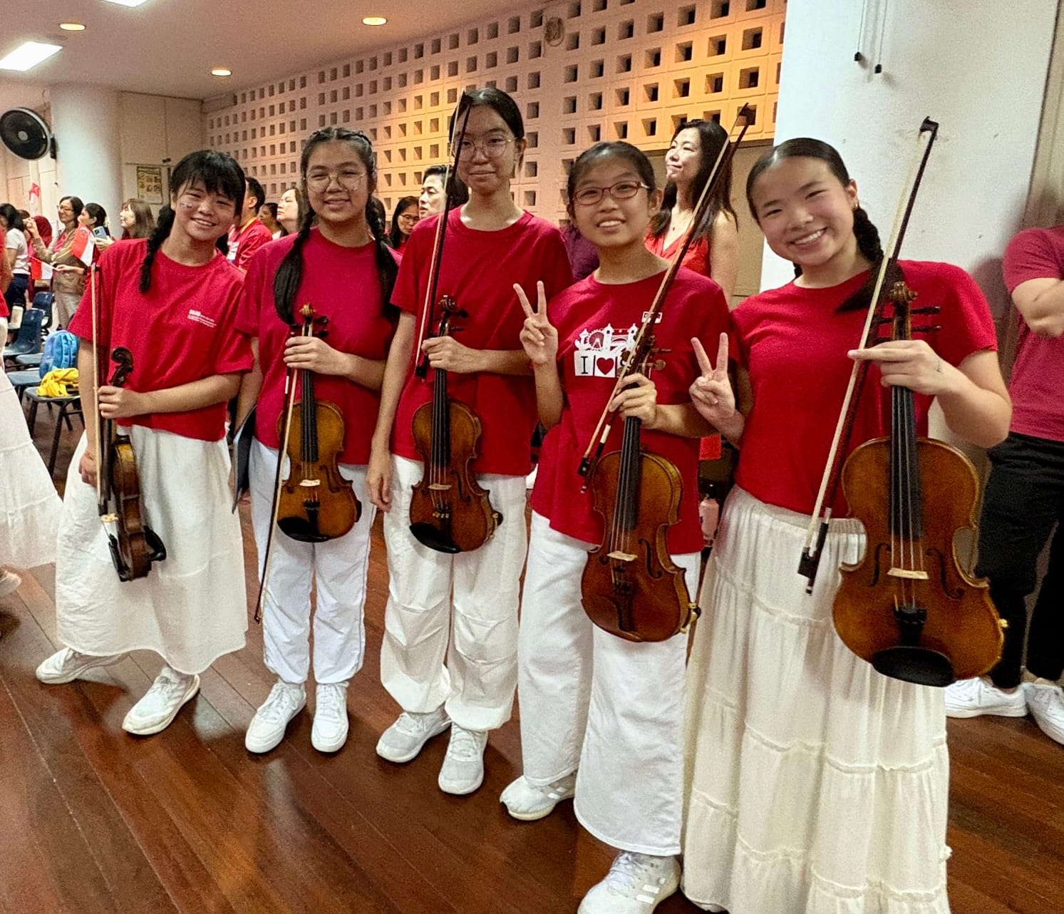 Five young girls in red shirts and white bottoms hold violins and smile, some making peace signs.