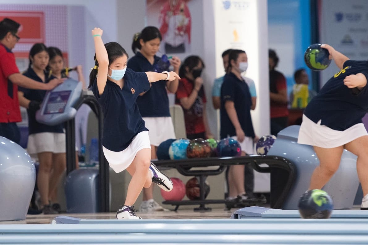 A young girl wearing a face mask celebrates after bowling, with other children and bowling balls visible in the background.