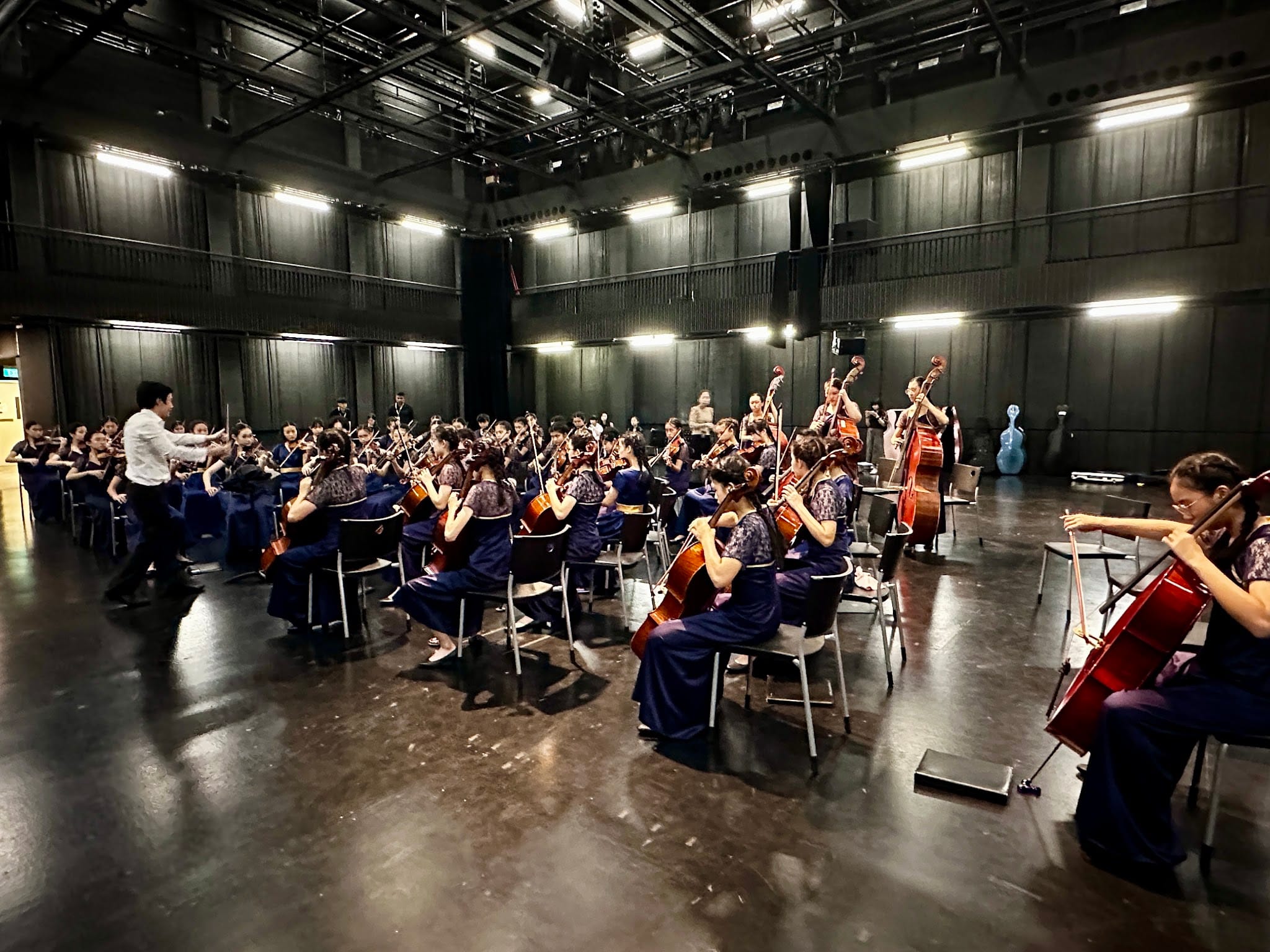 Large youth string orchestra in navy dresses rehearses indoors, led by a conductor in a white shirt.