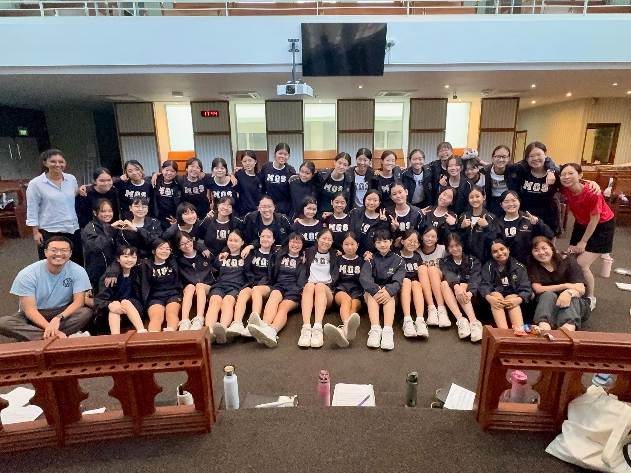 Large group of students in matching dark MGS uniforms posing together indoors with a few adults.