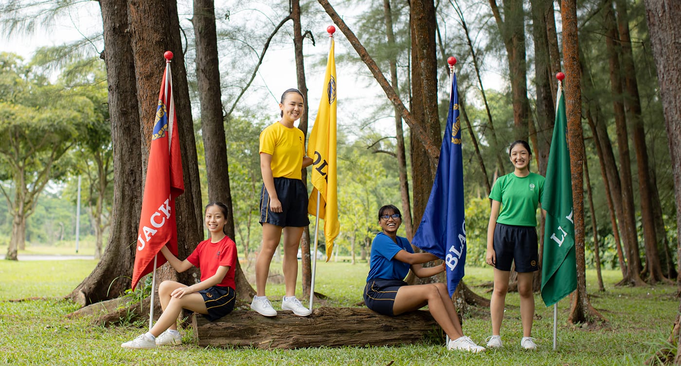 Four girls in red, yellow, blue, and green shirts pose with matching colored flags among tall trees on grass.
