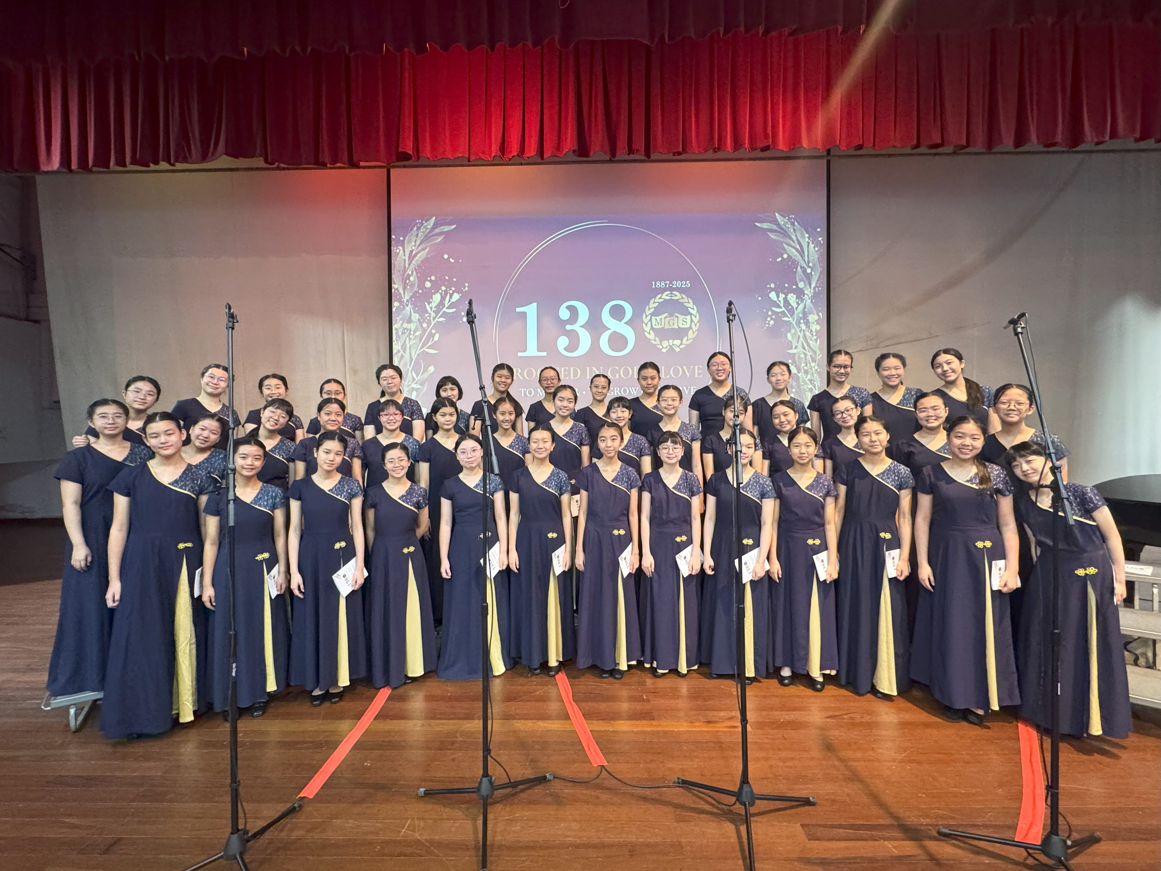 Large girls choir in navy dresses poses on stage before a screen showing "138" for a school anniversary event.