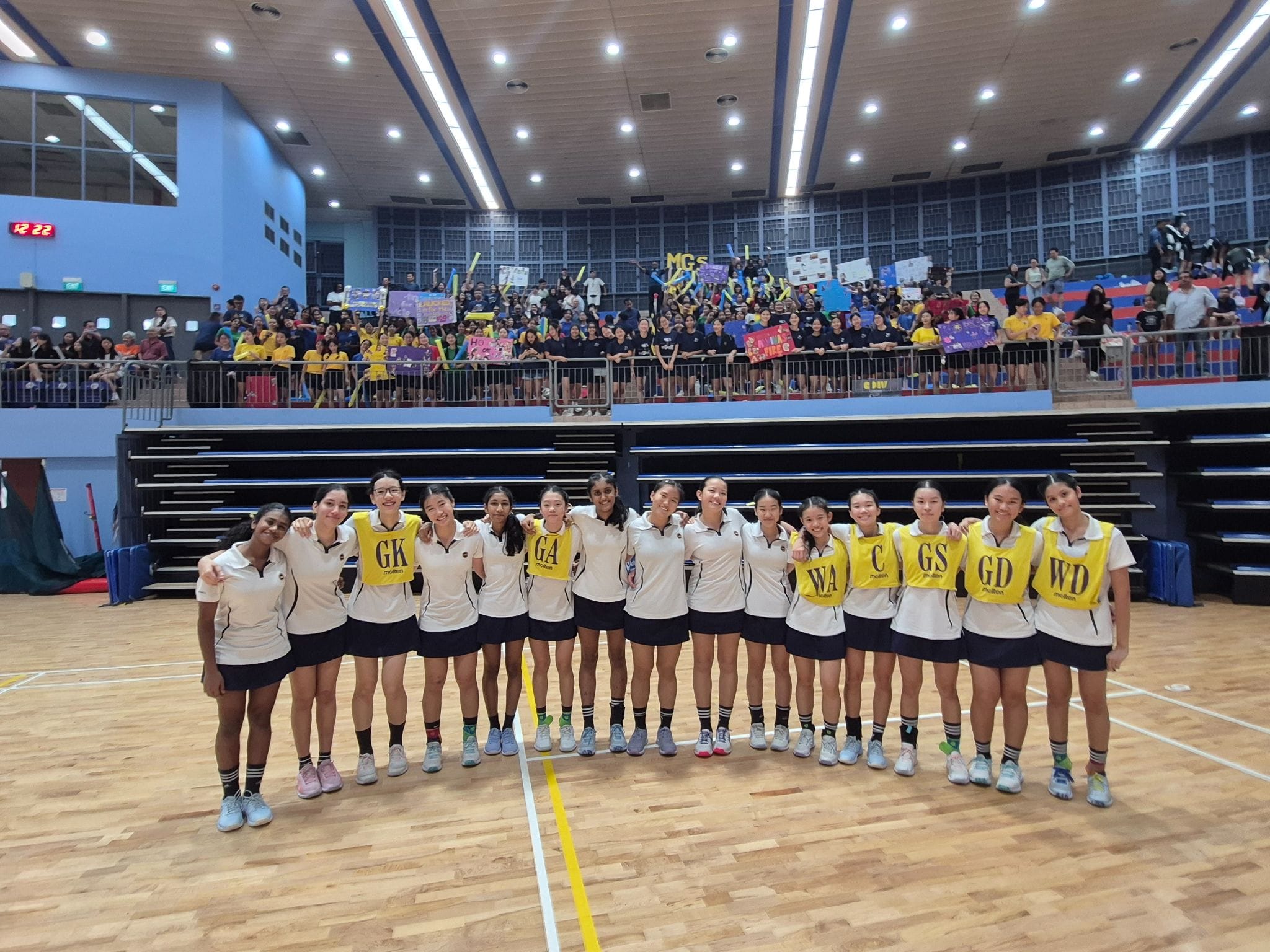 A netball team of teenage girls in white uniforms and yellow position bibs poses on a court with a packed crowd behind them.
